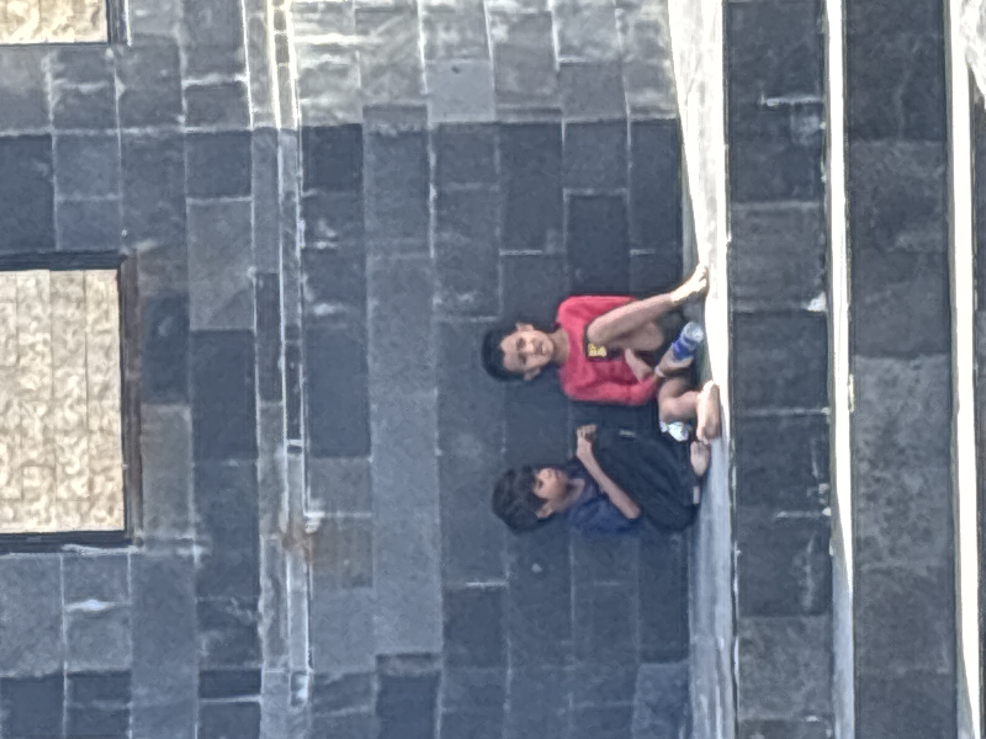 Two young boys, appearing to be of South Asian descent, are sitting on stone steps in front of a dark stone wall. The boy on the left is wearing a blue shirt and black pants, and is looking down at his hands. The boy on the right is wearing a red shirt and shorts, and is looking to his left. A blue plastic water bottle is on the step in front of the boy on the right. The wall appears to be part of a building or monument. The area is outdoors and appears to be sunny. The location is the Taman Ayun Temple in Mengwi, Indonesia.