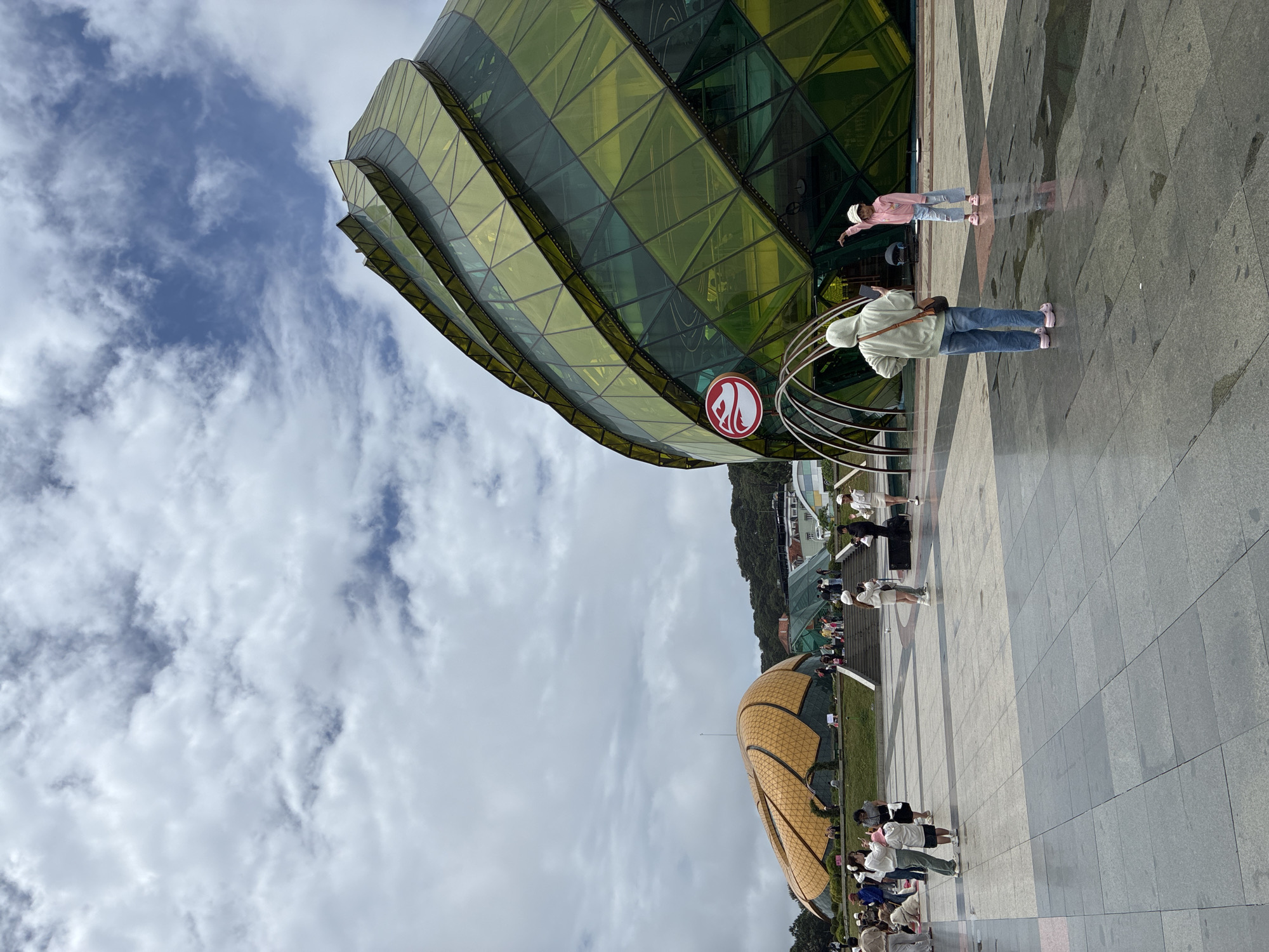 The image depicts the Da Lat Flower Park, a prominent tourist attraction in Da Lat, Vietnam, under a partly cloudy sky. In the foreground, a paved plaza is visible, with several people scattered about. Two individuals are positioned centrally: an adult wearing a light green hooded sweatshirt and blue jeans, holding a phone, and a child in a pink jacket and blue jeans, pointing towards the right. To the left, a cluster of people are gathered, some sitting and others standing, with one person taking a photograph.
Dominating the right side of the frame is a large, modern building with a distinctive, faceted green glass exterior, resembling a blooming flower. A red and white circular logo is affixed to its façade. To the left of this building, a series of metallic arches form a decorative entrance.
Further in the background, another architectural structure is visible, shaped like a large, golden-brown dome with segmented panels, also evoking a floral form. This dome is situated on a slightly elevated grassy area, accessible by a set of stairs where more people are seen ascending. The landscape beyond the structures features trees and buildings, suggesting an urban setting within a hilly terrain. The ground in the plaza shows wet patches, indicating recent rain.