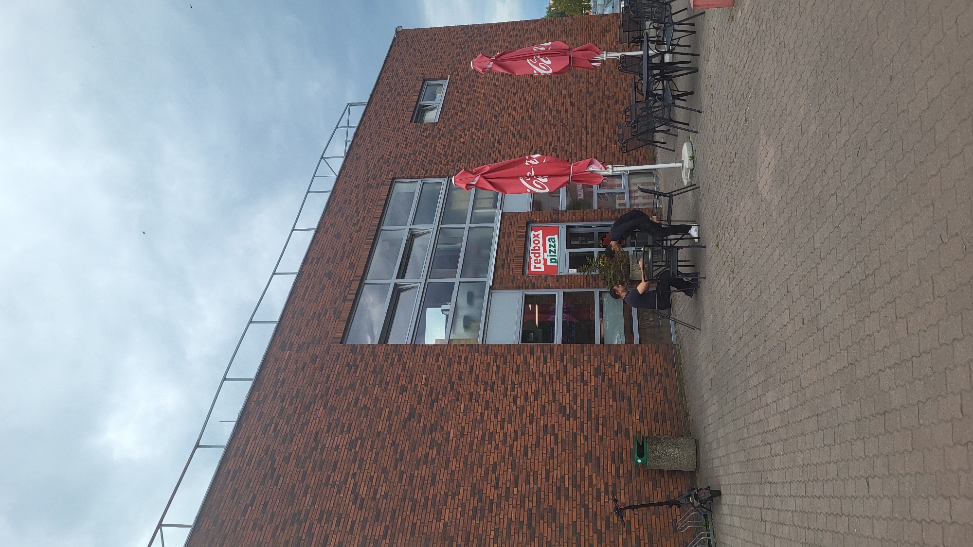 The image shows the exterior of a Redbox Pizza restaurant in Prešov, Slovakia, situated in a commercial area with a red brick facade and large windows. In front of the restaurant, there is an outdoor seating area with tables and chairs, partially shaded by large red umbrellas. Two individuals are seated at one of the tables, engaged in conversation. To the left, an electric scooter is parked next to a bicycle rack and a trash bin. The ground is paved with interlocking concrete blocks. The sky is overcast, suggesting a cloudy day.