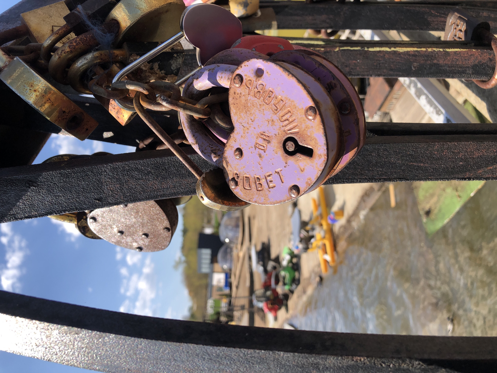 A close-up shot taken at an eye-level angle reveals a collection of love locks secured to a metal railing, with a blurred backdrop of the Votkinsk waterfront in Russia.
The primary focus is on several padlocks attached to the railing and linked together by a rusty chain. The locks exhibit signs of age and weathering, with a prominent, heart-shaped purple padlock in the foreground. Cyrillic inscriptions are visible on the purple lock.
The background provides a glimpse of the waterfront area, featuring watercraft, possible recreational structures, and sparse vegetation along the water's edge. The overall scene suggests a location where individuals have chosen to commemorate their relationships by placing love locks, a common practice in many cities worldwide.