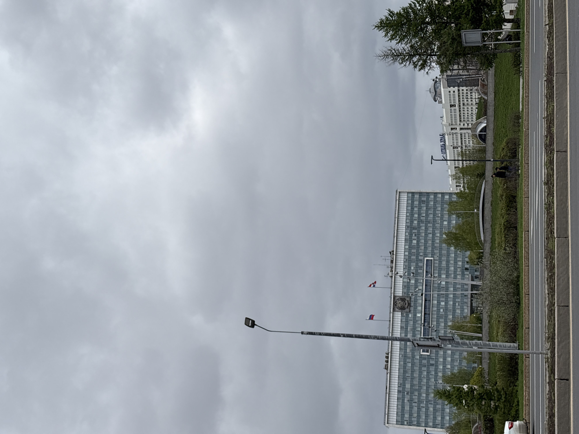 The image depicts a street scene in Perm, Russia. A large, multi-story building with numerous windows dominates the right side of the frame. Two flags, likely Russian, are visible on the building's roof. A tall lamppost stands in the foreground, slightly obscuring the view. A roadway runs along the right edge of the image, adjacent to a strip of green space with trees. Further in the background, another building is visible, with the signage "ОТЕЛЬ УРАЛ" ("Hotel Ural") displayed. The sky is overcast with grey clouds. Two individuals are discernible in the green space near the "Hotel Ural" building.