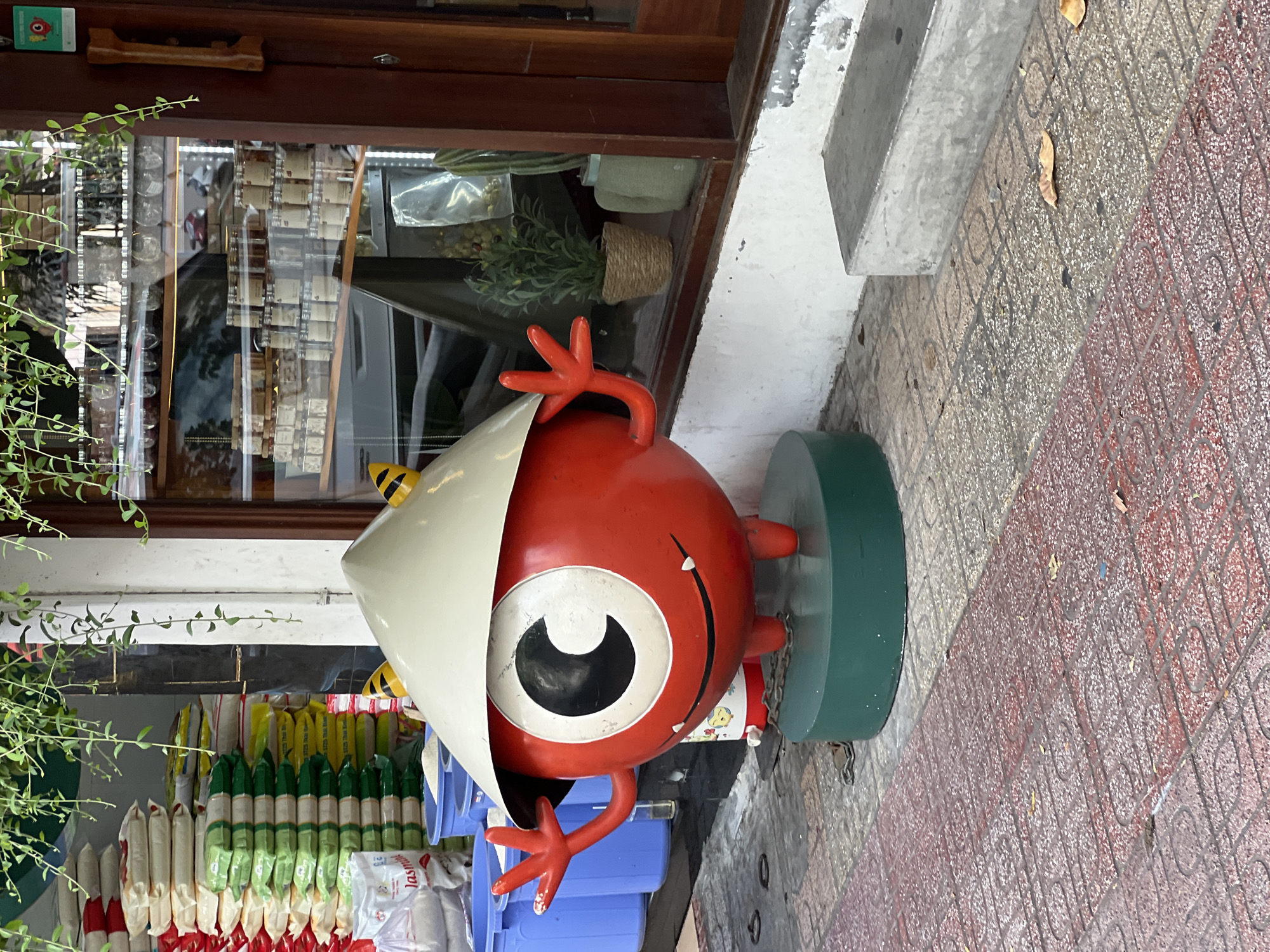 In Nha Trang, Vietnam, a vibrant red, cartoonish character stands on a green circular base. The character, with a large white eye and a conical hat, wears a cheerful expression and has its left hand raised in a wave. The scene takes place outdoors, with a store window featuring shelves of jars and plants in the background. Piles of rice bags are stacked to the left of the character, and blue buckets are visible in front of them. The ground is paved with red and grey tiles, and a concrete bench is present to the right. The overall impression is one of a lively street scene in a Vietnamese city.
