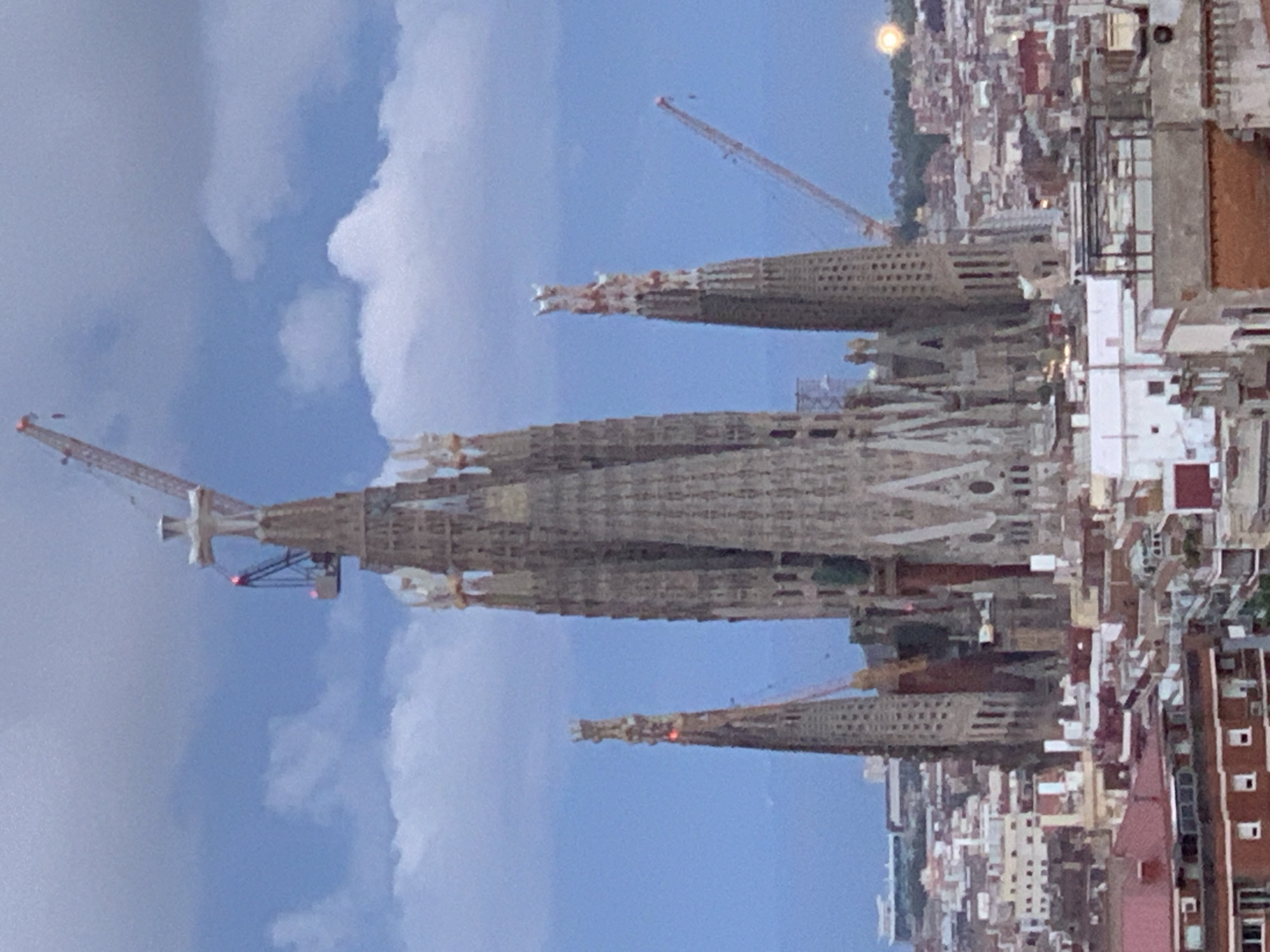 The image shows a view of the Sagrada Familia in Barcelona, Spain. The basilica's spires are prominent, with construction cranes visible around the building. The sky is blue with some clouds. Residential buildings are seen in the background.