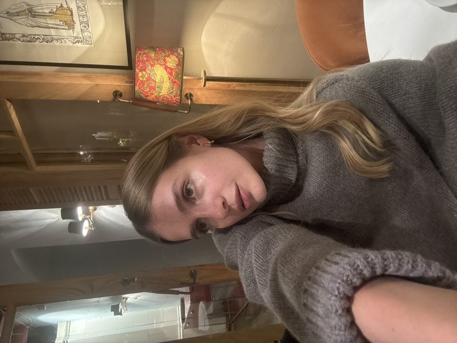 A young adult female is positioned in an indoor setting, specifically identified as the PurPur cafe/restaurant on Betlemi Street in Tbilisi, Georgia. She has shoulder-length blonde hair and fair complexion, wearing a thick, grey turtleneck sweater. Her left arm is partially visible, with her hand out of frame, suggesting a selfie pose or arm resting. She is gazing upwards and to her right.
To the subject's left, a large mirror reflects a section of the establishment's interior, showing a wall-mounted brass light fixture with two black conical shades. Further into the reflection, a light-colored wall and red-upholstered chairs around a table are discernible. Wooden architectural elements, possibly window frames or dividers with louvered sections, are also reflected.
To the subject's right, a wooden panel or door frame is visible. A distinctive wall lamp is attached, featuring a red fabric lampshade patterned with yellow birds and foliage. Above this lamp, a framed artwork depicts a standing robed figure. The edge of an orange-brown upholstered chair is present in the mid-right, and a white table surface is visible in the lower right foreground. The ambient lighting is artificial and dim.