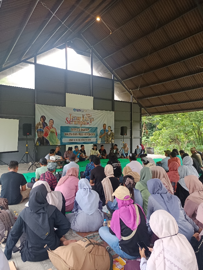 The image depicts an indoor event in Mayang, Indonesia, within a large, open-sided building featuring a high, corrugated metal roof supported by dark metal trusses. The walls are dark grey panels, and the floor is concrete, partially covered with green and patterned mats. To the right, the structure is open, revealing lush green trees and foliage.
In the foreground and midground, a diverse audience of approximately 50-70 individuals is seated on the floor, facing forward. The attendees are casually dressed; many women wear hijabs in various colors (e.g., black, light blue, pink, beige), while men wear shirts and trousers. Several individuals are observing the front, some are looking down at their phones, and personal bags and water bottles are scattered around them on the floor.
At the front of the venue, approximately 8-10 panelists or presenters are seated cross-legged on a raised green mat. They are casually dressed, some in patterned shirts, others in plain attire. Microphones are present for communication. A white projector sits on the floor in front of them. Behind the panelists, a large white banner is displayed, reading "Temu Karya Serumpung 2023" (translating to "Cluster Work Meeting 2023") at the top, followed by "Semesta Ingatan: Trauma dan Imaji Kebebasan" (Universe of Memory: Trauma and Images of Freedom), and "Jumat, 25 & 26 Oktober 2023" (Friday, October 25 & 26, 2023) at the bottom. The banner also features illustrative portraits of individuals in traditional and contemporary Indonesian clothing.
Flanking the stage area are two black PA speakers mounted on stands. To the left of the stage, a white projection screen on a tripod stand is visible. The scene is illuminated by ample natural daylight entering from the open sides and gaps in the structure, supplemented by a single visible overhead light fixture in the ceiling.