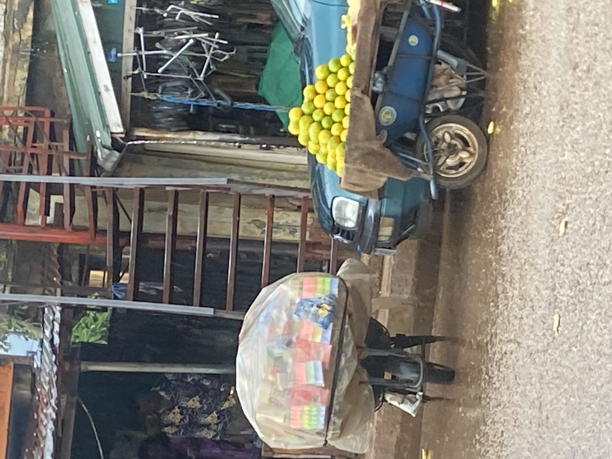 This image shows a street scene in Bukuru, Nigeria. The primary subjects are a blue tricycle carrying a load of yellow fruits, likely lemons, and another tricycle covered with a clear plastic sheet containing various colorful items, possibly snacks or small packaged goods. The background includes structures with metal scaffolding, some covered with green sheeting, and stacks of what appear to be industrial materials like pipes and metal frames. The ground is covered with gravel.
The scene suggests a commercial or market environment, with the tricycles likely being used for vending or transportation of goods. The presence of building materials and scaffolding hints at ongoing construction or a storage area adjacent to the street. The lighting appears to be natural daylight, suggesting the photo was taken during the day. The overall impression is one of a busy, practical, and possibly informal commercial setting. No people are clearly visible in the frame, though the presence of the goods implies human activity. There is no visible text in the image.