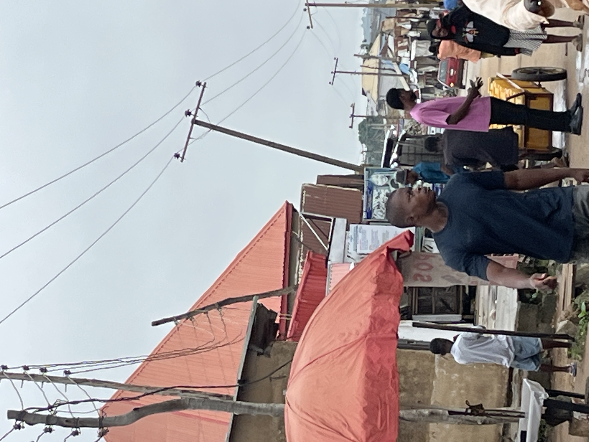 The image depicts a street scene in Abuja, Nigeria, under an overcast sky. In the foreground, a man in a dark blue t-shirt walks towards the right. To his left, a person in a white t-shirt and blue shorts stands under a large, orange umbrella, facing away from the camera. Behind them, a red-tiled roof of a building is visible. The street is lined with utility poles and wires crisscrossing the sky. Further down the street, several people are seen, including a man in a pink t-shirt, a woman in a black hooded jacket with a bee design, and others partially obscured. A red car is parked on the right side of the street, and various market stalls or makeshift structures are present. The ground appears to be paved with dirt and some debris.