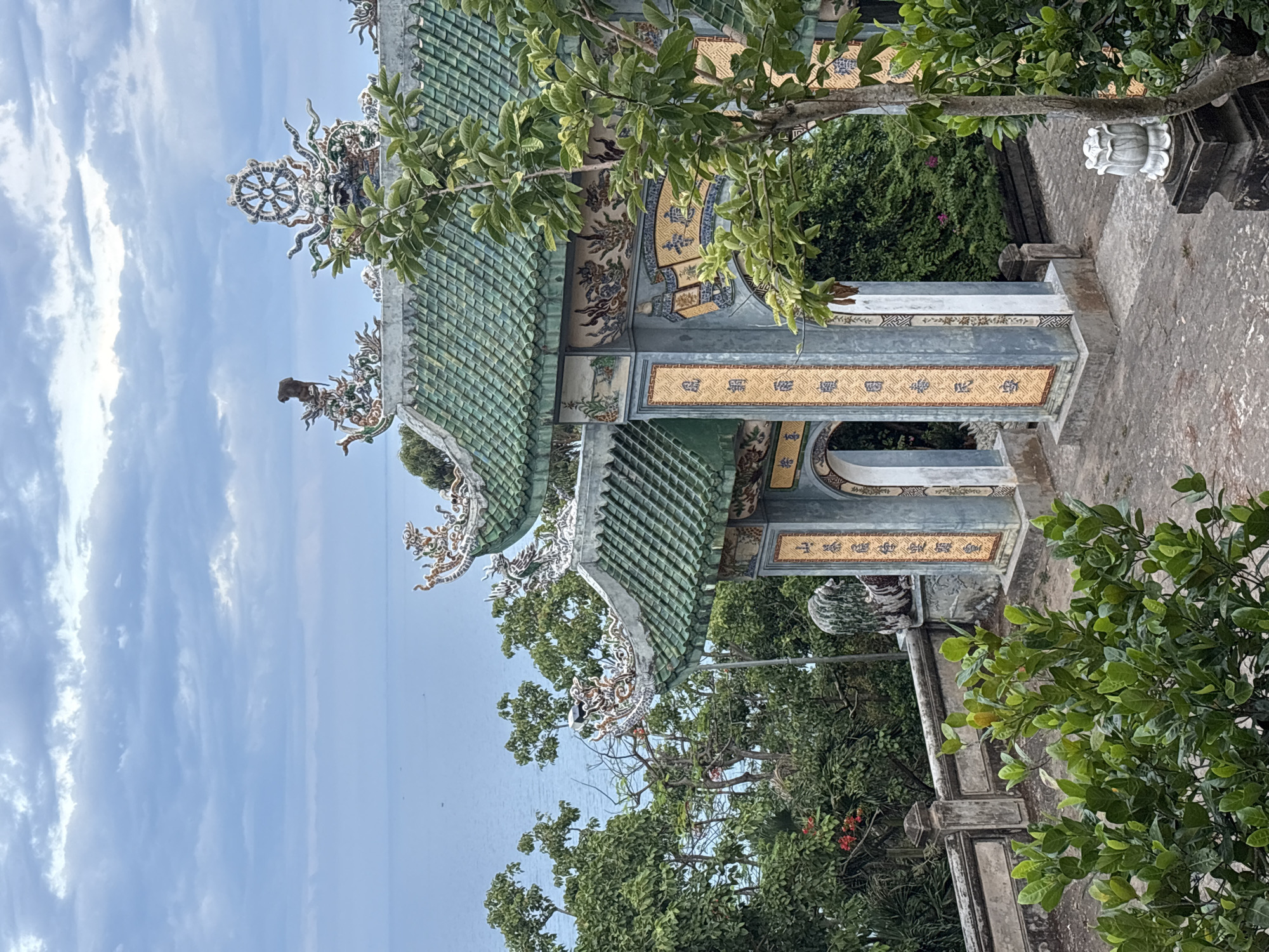 This is an exterior, eye-level shot of the entrance gate to the Linh Ung Pagoda in Da Nang, Vietnam. The gate features ornate, green tiled roofs with dragon motifs. The sky is cloudy and the ocean is visible in the background. Lush green trees surround the pagoda entrance. A stone lion statue is visible on the left side of the gate. The gate itself is made of gray stone and decorated with colorful tile panels featuring Asian calligraphy.