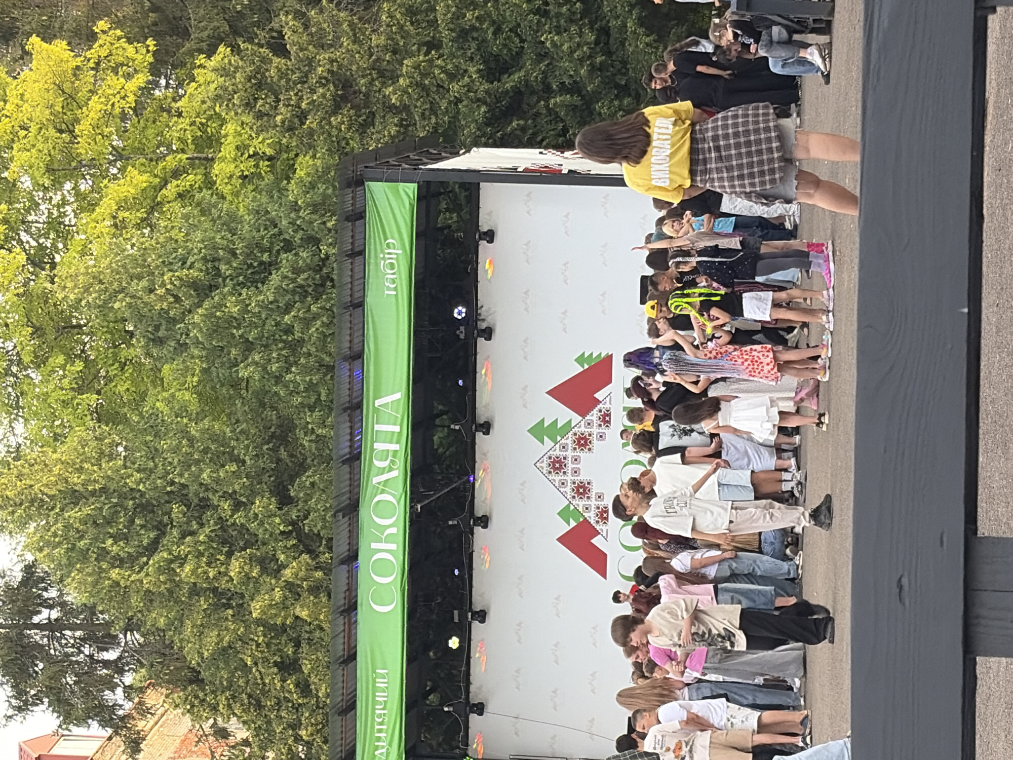 The image shows a crowd of people, mostly young, gathered in front of a stage. A banner with Ukrainian text is displayed above the stage. Some people are dancing and interacting, while others appear to be observing. The environment suggests an outdoor event, possibly a festival or concert, with trees and foliage visible in the background. The lighting suggests it is daytime, possibly late afternoon.