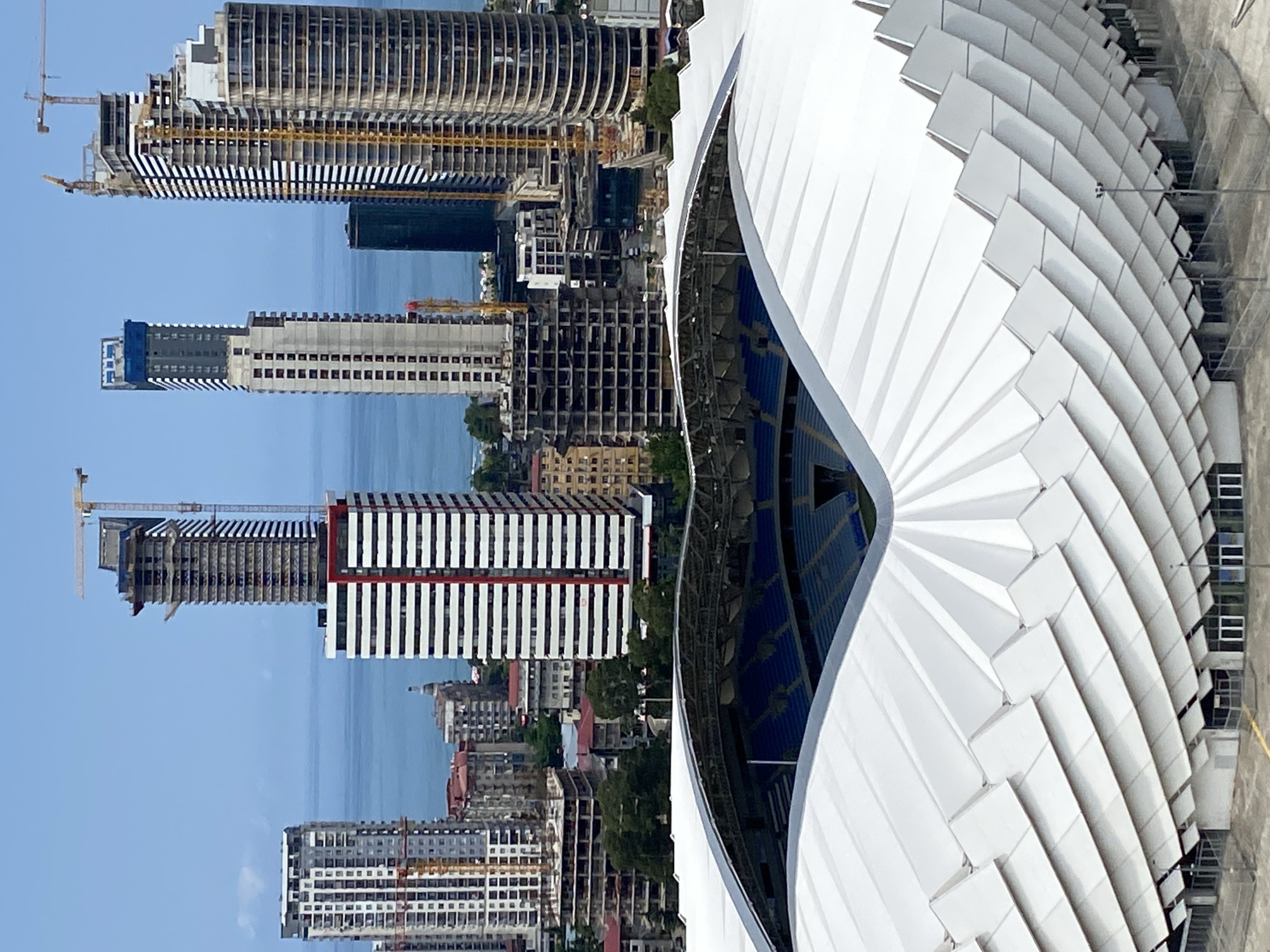 This is an aerial, high-angle, vertical, medium shot of the Batumi Stadium in Batumi, Georgia. The stadium's white, pleated roof is in the foreground, with rows of blue seats visible inside the stadium. Beyond the stadium, a cityscape of tall buildings, many of which appear to be under construction, stretches towards the Black Sea, which is visible in the background. Several cranes are present amongst the buildings, indicating ongoing construction. The sky is a clear, pale blue.