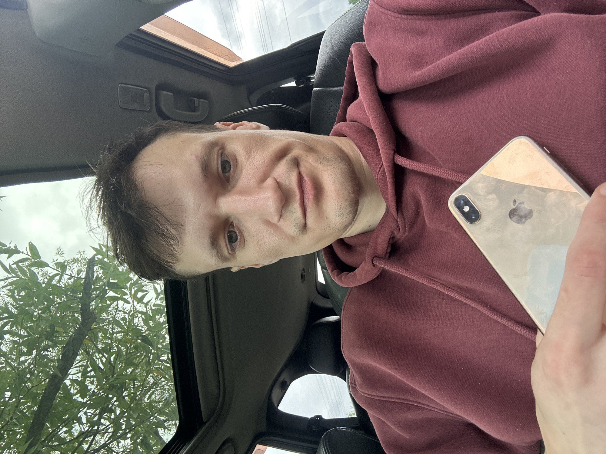 A young man with short brown hair and fair skin is sitting in the driver's seat of a car, taking a selfie. He is wearing a maroon hoodie. The car has a sunroof open, revealing green foliage and a cloudy sky above. The man is holding a gold-colored smartphone in his right hand, which is partially obscuring his lower face. The interior of the car is visible, including black leather seats and the car's headliner. The image appears to be taken in an outdoor setting, possibly in Makiv, Ukraine, based on the surrounding greenery.