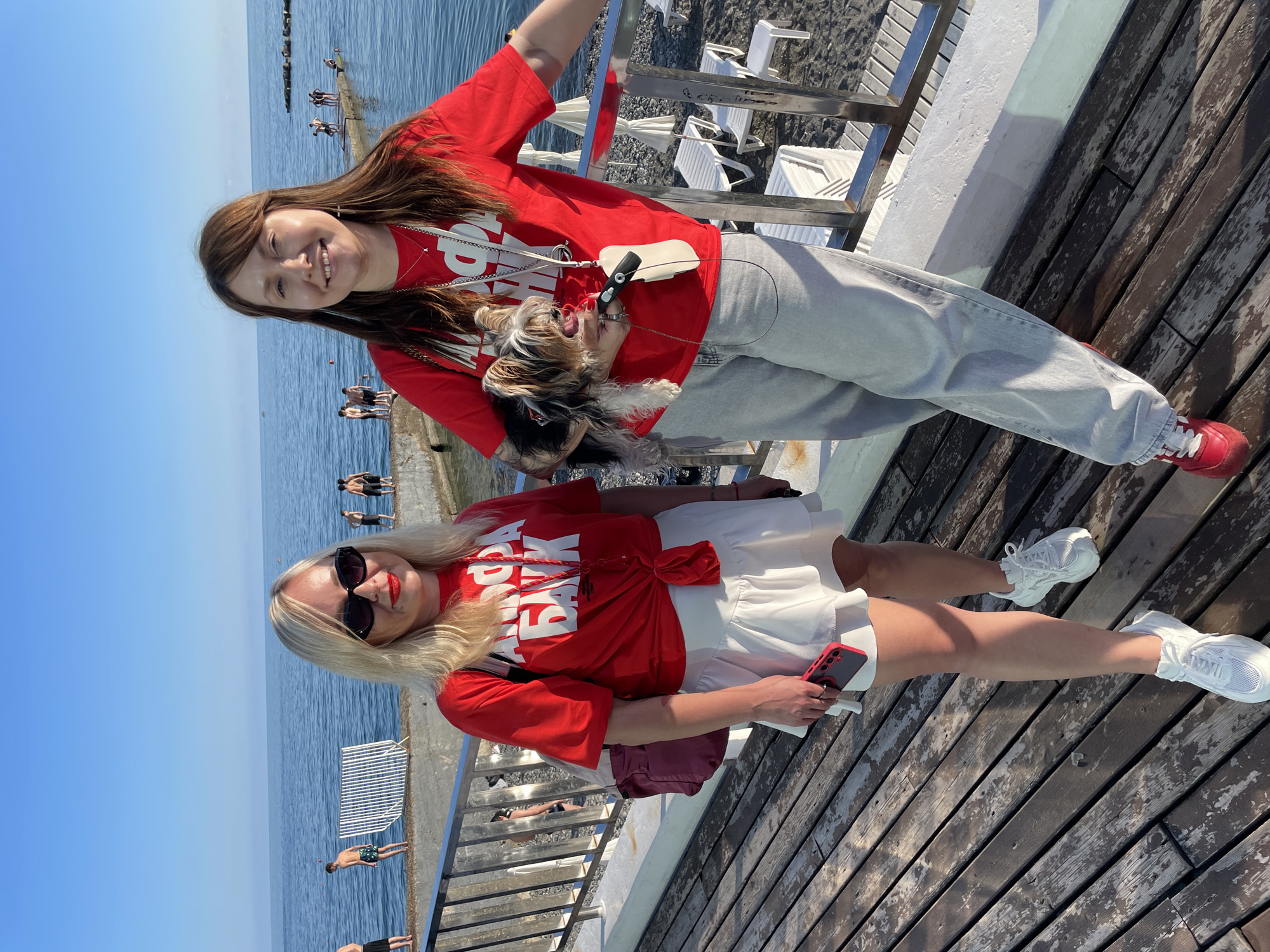 Two women and a small dog are standing on a wooden boardwalk next to a body of water, likely the Black Sea, on a sunny day. The woman on the left has blonde hair, sunglasses, and is wearing a red t-shirt with white lettering and a white ruffled skirt. The woman on the right has brown hair and is holding a small dog, wearing a red t-shirt and light-colored pants. In the background, several people are visible near the water, and there are lounge chairs and umbrellas on a pebble beach. The setting appears to be a resort area, possibly in Sochi, Russia, given the architectural style of the railing and the presence of lounge chairs common in such locations.