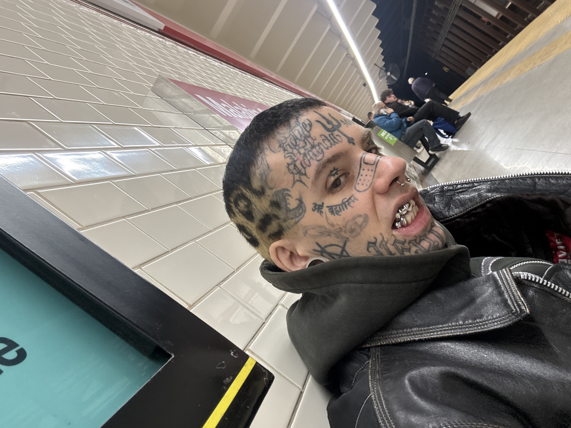 A low-angle medium shot captures a tattooed individual wearing grillz and a black leather jacket in what appears to be a subway station. The person is leaning against a wall tiled with white rectangular tiles. In the background, people are seated on benches along the platform. A black metal frame with a blue screen is visible on the left side of the frame. The location could be a subway station in Buenos Aires, Argentina.