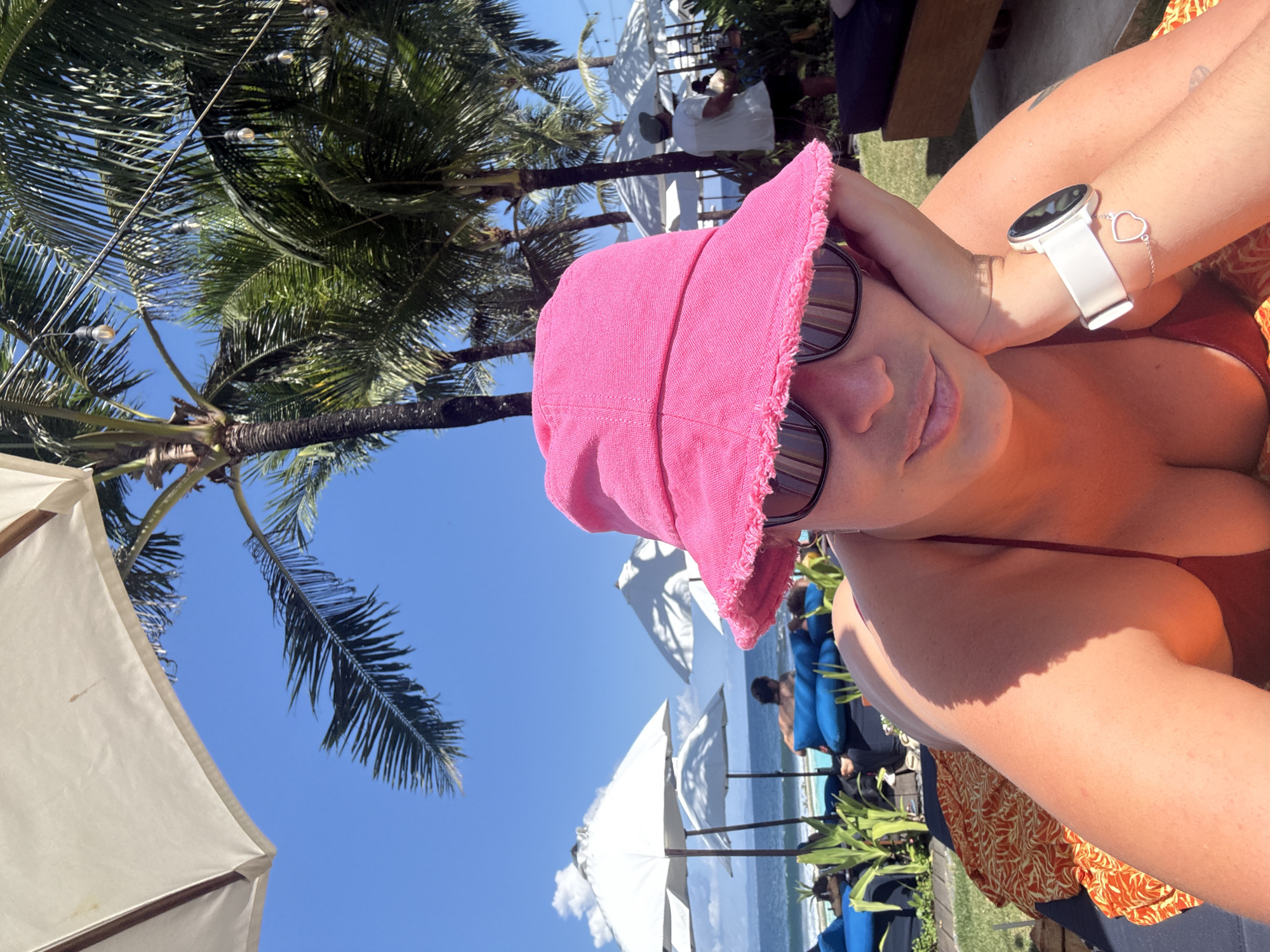 A selfie captures a woman in a pink hat and sunglasses on a sunny day at a beach in Medahan, Indonesia. She is wearing a white smartwatch with a heart bracelet on her wrist and a red swimsuit. Behind her, the clear blue sky is dotted with white beach umbrellas. Palm trees frame the scene. In the background, people are visible on lounge chairs near the ocean. A white umbrella and vegetation are also present.