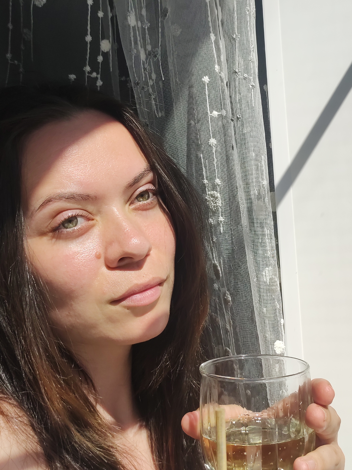 A woman with fair skin and dark hair holds a glass containing a light-colored liquid, possibly wine, in her right hand. Her green eyes are directed slightly upward and to the right. She stands near a window covered with a white, floral patterned curtain. Sunlight casts shadows across her face, the curtain, and the white wall beside the window. The scene's location in the city of Фоки, Russia is unsubstantiated.