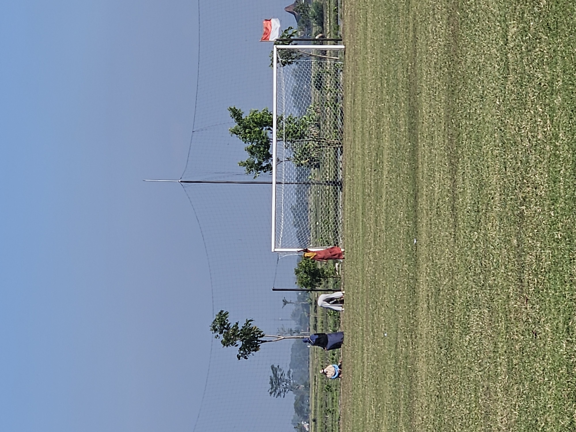 The image shows a soccer field in the city of Perak, Indonesia. The field is green and well-maintained. There is a soccer goal at one end of the field. In the background, there are some trees and a flag of Indonesia. There are four people in the image, all wearing traditional Indonesian clothing. One person is sitting on the ground, and the other three are standing. The people appear to be engaged in some sort of activity, but their exact actions are unclear. The sky is clear and blue.