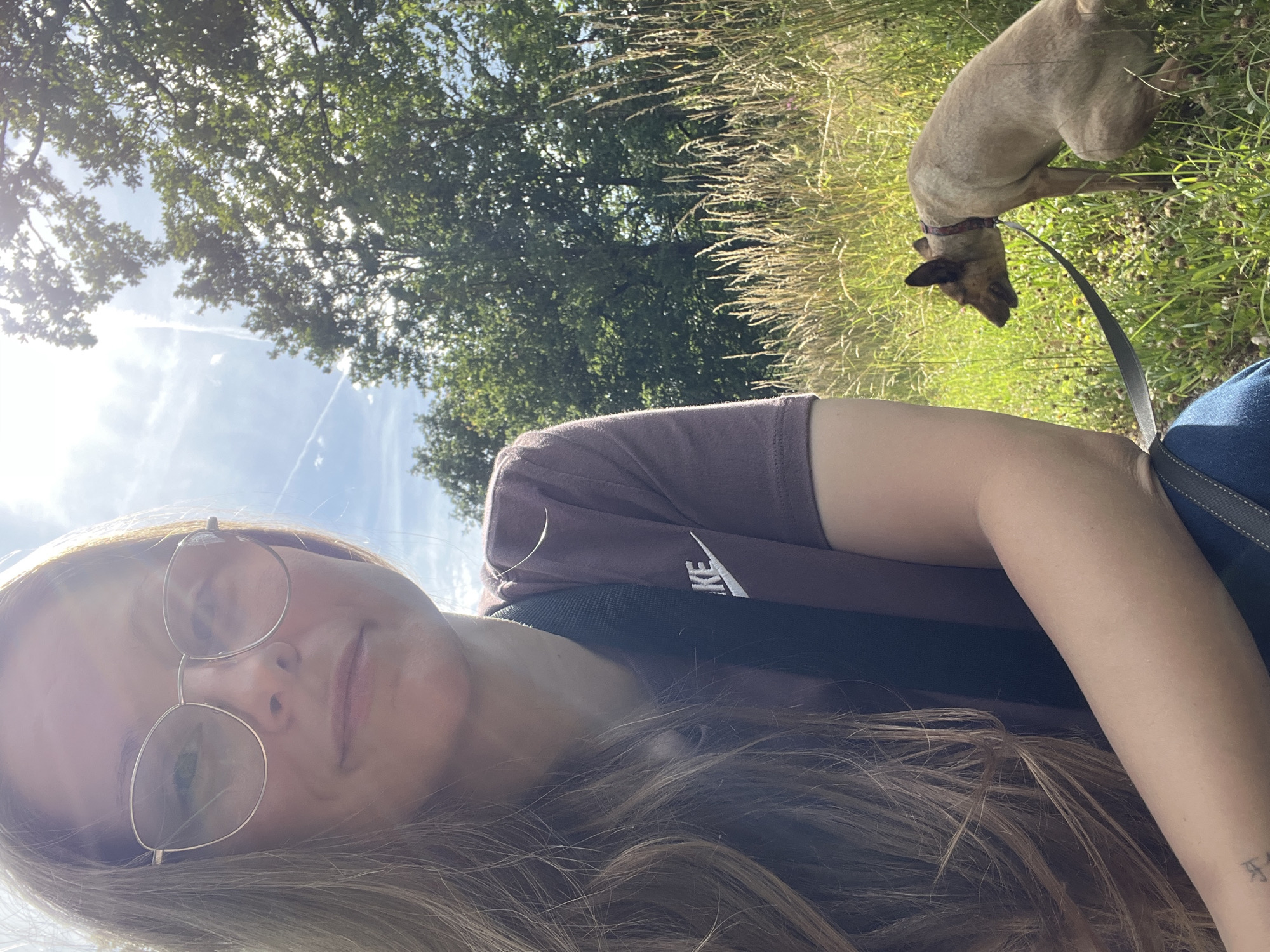 The image shows a woman taking a selfie outdoors, with her dog in the background. The woman is wearing glasses and a maroon t-shirt with a white logo. She has long, blonde hair that covers part of her face and arm. The dog, which appears to be a light brown color, is leashed and standing in tall grass. Behind them, trees and a bright sky with some wispy clouds are visible. The sun is shining, suggesting it is daytime. The scene appears to be a park or a natural area, possibly in Saint Vith, Belgium. The woman's expression is relaxed and happy, and the overall mood is pleasant and summery. There is visible text on the woman's t-shirt, appearing to be a white logo on the right side of her chest.