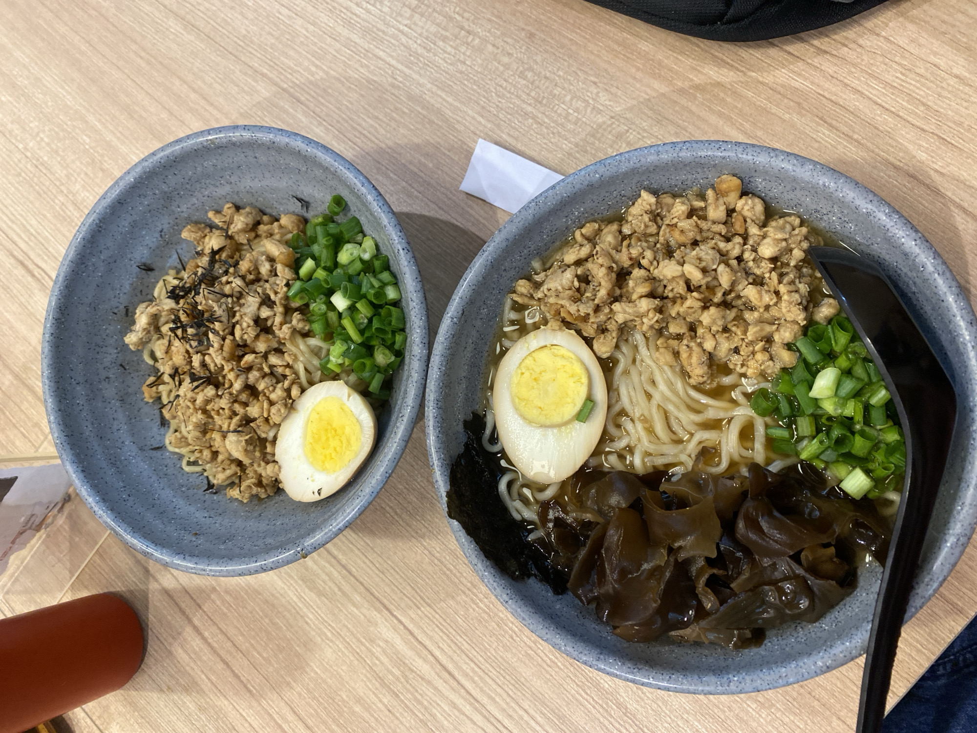 Two bowls of ramen are on a light wooden table. The bowl in the foreground contains noodles, ground meat, sliced green onions, a half-boiled egg, wood ear mushrooms, and a piece of nori. The bowl in the background contains noodles, ground meat, sliced green onions, and a half-boiled egg. A red thermos is visible to the left of the bowls, and a black bag is partially visible to the right. The scene appears to be in a restaurant in Banjarbaru, Indonesia, based on the typical presentation of ramen and the wooden table setting common in Indonesian eateries.
