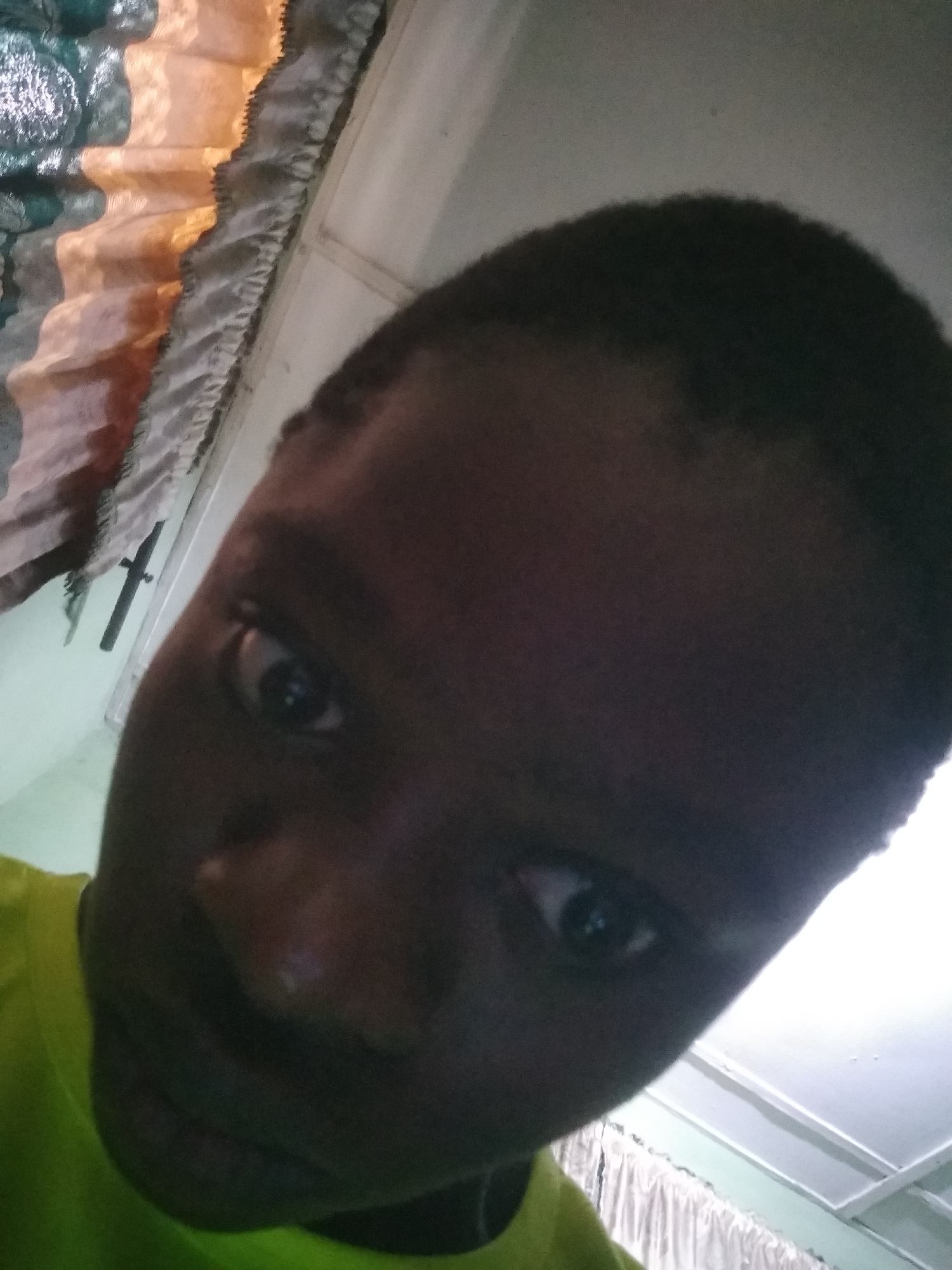 An upward-angled selfie of a dark-skinned person with short, dark hair. The person is wearing a bright yellow shirt. Behind the person, a curtain hangs along the top edge of the frame, and a white ceiling is visible. A cross-shaped object is affixed to a wall behind the person. The location appears to be indoors. There is no definitive information to ascertain this image was taken in Shangisha, Nigeria.