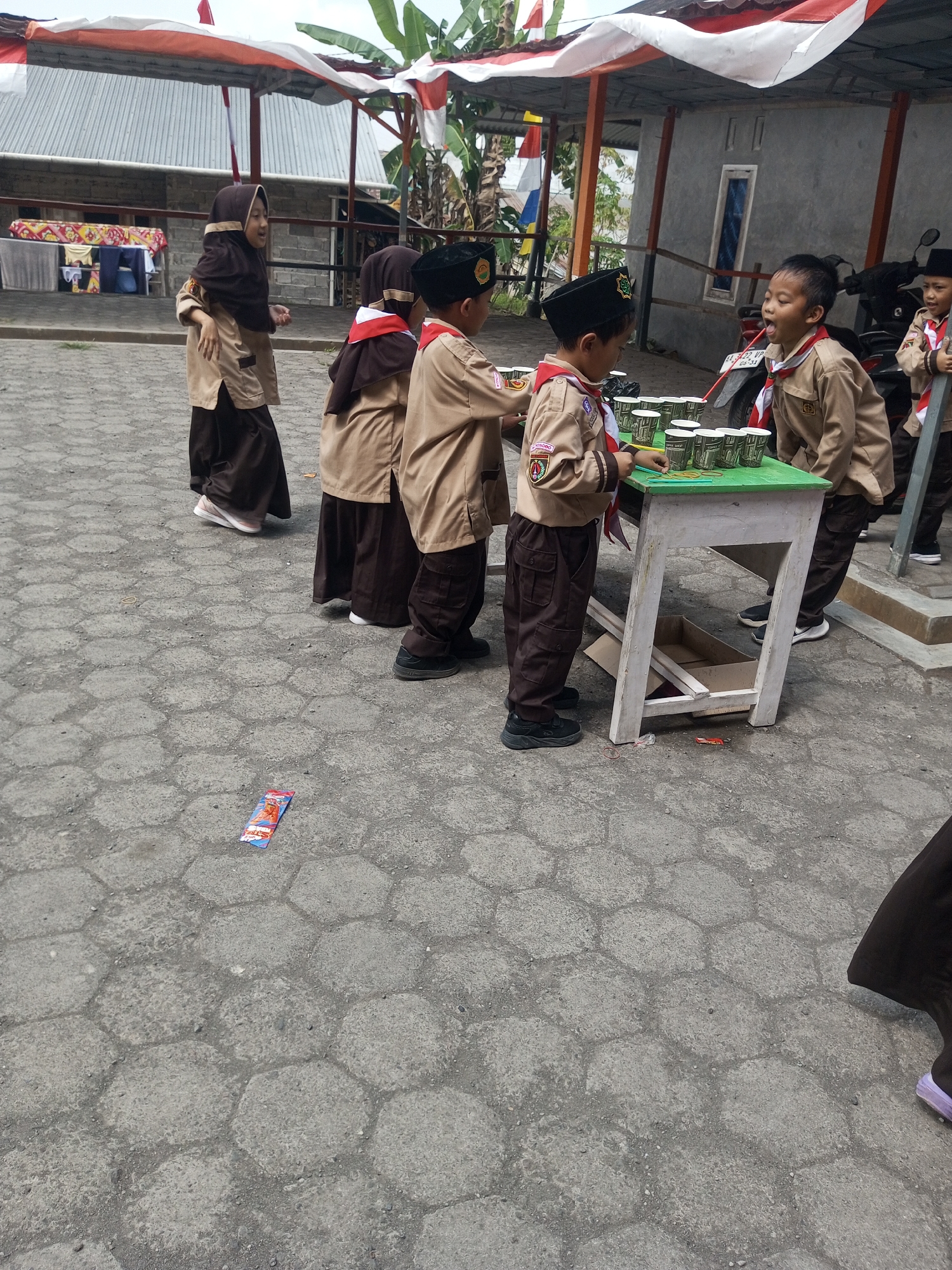 In the city of Kalibeber, Indonesia, a group of children in scouting uniforms are gathered around a green-topped table. The children, both boys and girls, are wearing brown shirts, dark brown pants, and some are wearing dark headscarves or black hats with insignias. They are actively engaged with a game involving numerous small cups arranged on the table, with some children holding what appear to be rubber bands. The table itself is white and has a wooden structure underneath, with a cardboard box visible below. The ground is paved with hexagonal concrete blocks. In the background, there are structures with corrugated roofs, decorative banners in red and white, and a motorcycle. A small, colorful wrapper lies discarded on the pavement in the foreground.