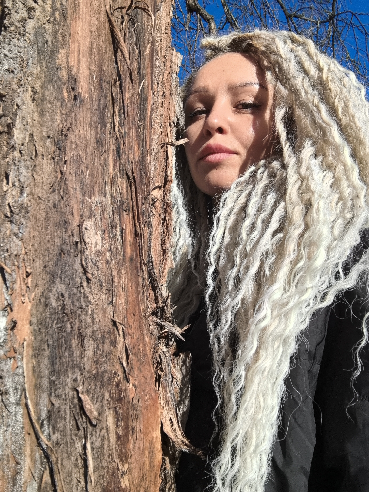 A medium-angle shot shows a woman with blonde dreadlocks standing next to a tree. The tree is in the left portion of the image. It has coarse bark. The woman's face is partially in shadow. She is wearing a black jacket. The sky is visible in the upper right corner of the image. Bare tree branches are also visible in that area.
While the instructions mention Noviy, Russia, this information cannot be verified by visual features in the image.