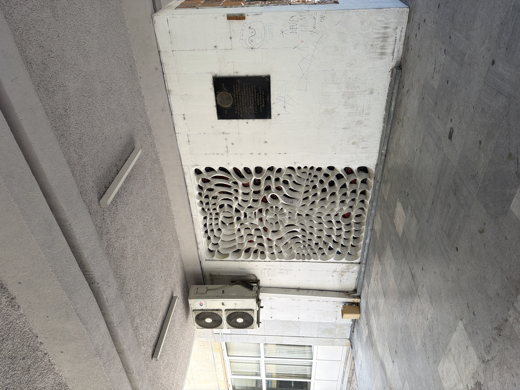 The image captures an exterior view of a building with a textured concrete ceiling supported by a beam. Two fluorescent light fixtures are visible on the ceiling. Below, a white wall features an air conditioning unit mounted on a concrete pillar to the left, with a glass-paneled door and window next to it. To the right of the air conditioning unit is a decorative white lattice artwork with a repeating organic pattern, revealing glimpses of red material behind it. Further to the right on the wall, a dark plaque is affixed, bearing text and an emblem. Below the plaque, there are various instances of graffiti, including drawings and text. A light-colored trash receptacle is placed on the ground near the base of the wall. The ground is paved with large, grey square tiles, some showing signs of wear and staining. The overall scene suggests an outdoor public space, possibly an entrance or walkway of a building in Pazardzhik, Bulgaria, given the presence of a historical plaque that likely commemorates an event or individual relevant to the city.