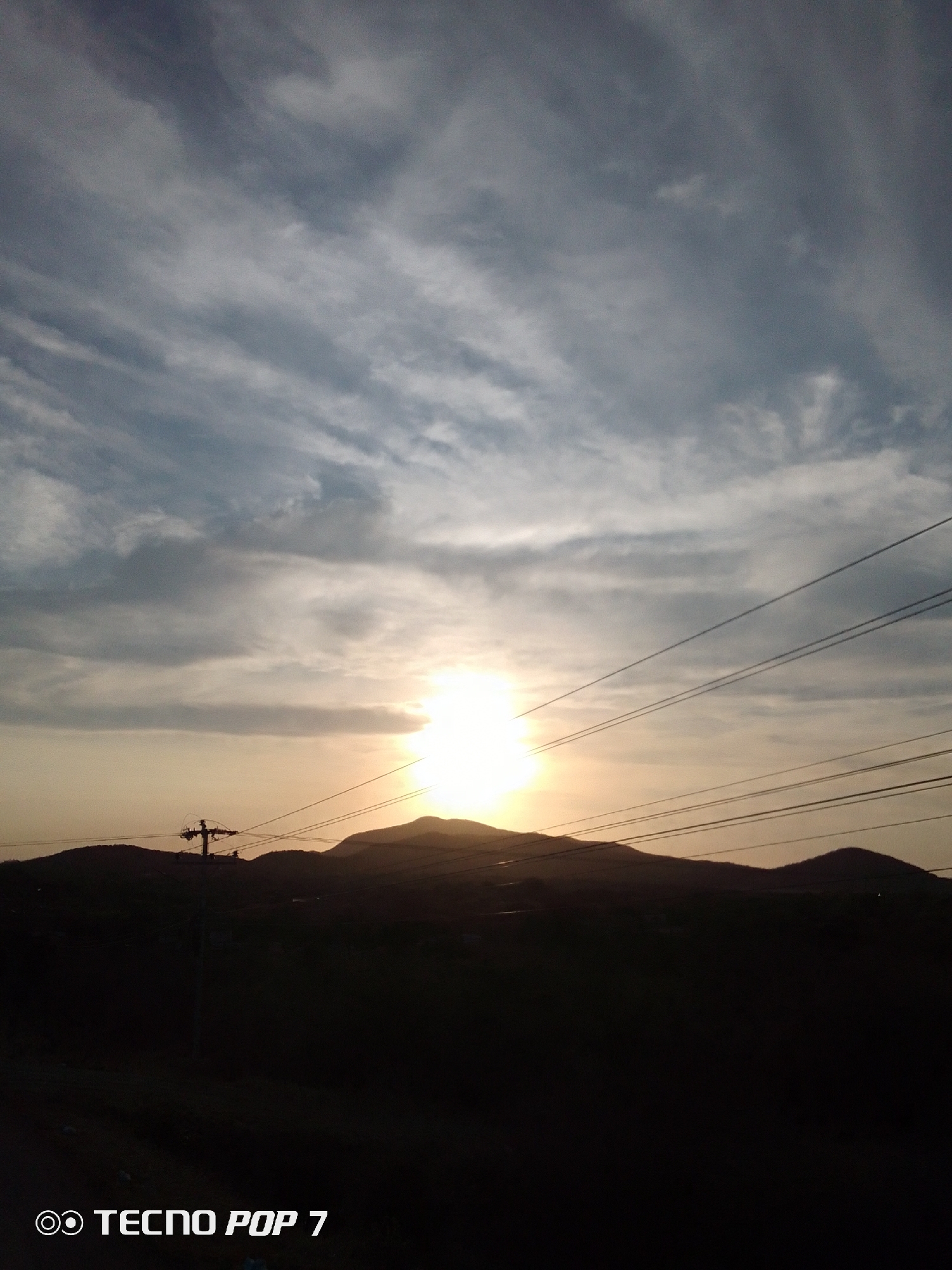 The photograph depicts a landscape in Altagracia, Venezuela, at sunset. Silhouetted mountains dominate the middle ground, with the sun setting directly behind the central peak. Power lines stretch across the sky. The sky displays streaks of clouds illuminated by the setting sun. The foreground is dark. A utility pole is visible on the left. The lower portion of the image shows the text "TECNO POP 7".