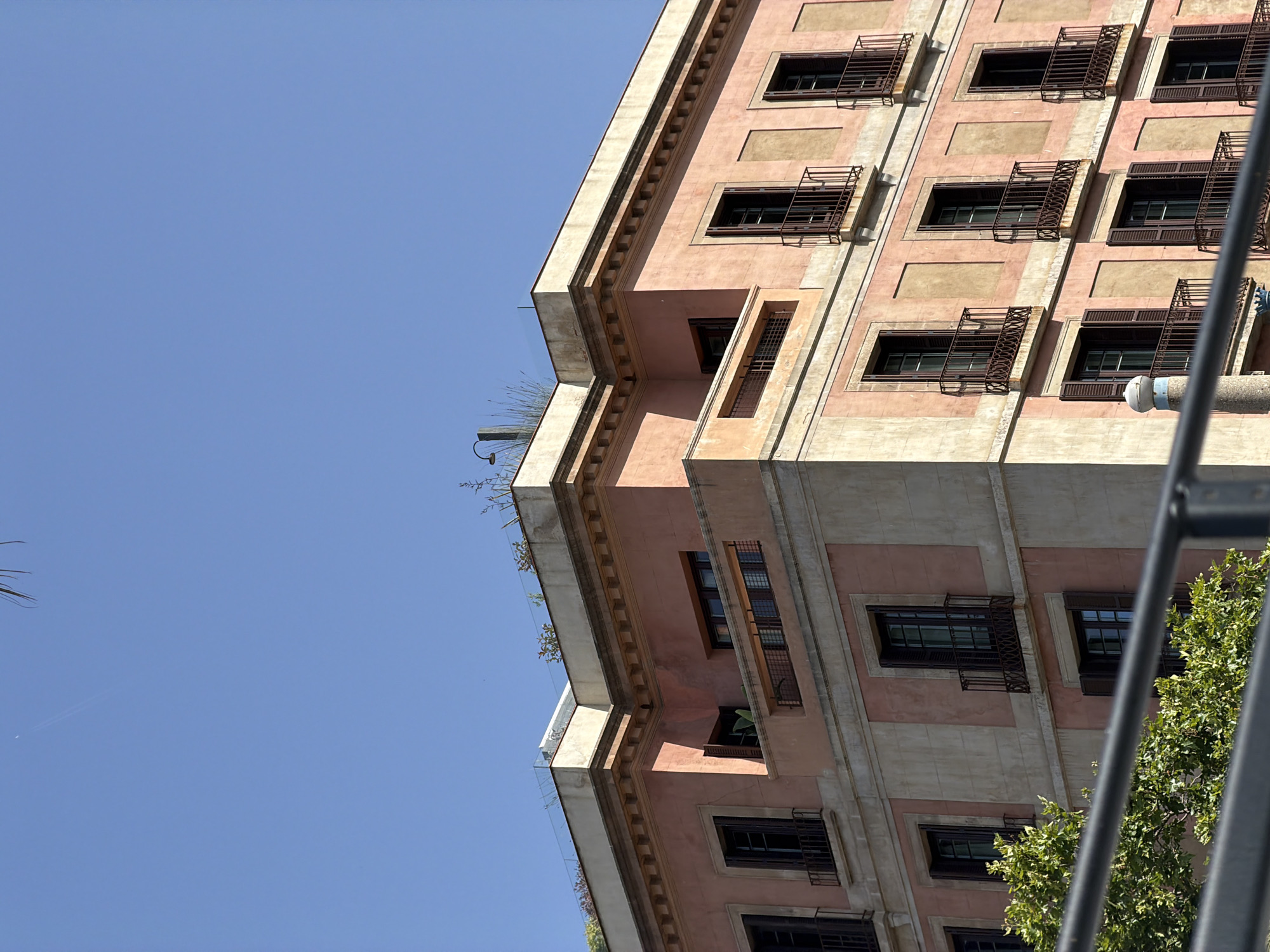 The image captures a close-up, angled view of the upper floors and roofline of a historic pink-hued building in Barcelona, Spain, against a clear blue sky. The architecture features multiple rows of windows with dark wooden shutters and wrought-iron balconies. Some windows are open, revealing glimpses of interiors, while others are closed. The building's facade is adorned with decorative moldings and alternating sections of light-colored stone and pink plaster. The rooftop reveals a modern glass barrier and sparse vegetation, suggesting a terrace or garden space. A tall, thin chimney-like structure with a rounded top is visible on the right side of the rooftop. In the foreground, out of focus, is a dark gray railing or fence, with the lush green foliage of a tree visible below it. The overall scene conveys a sense of urban life in a historic European city.