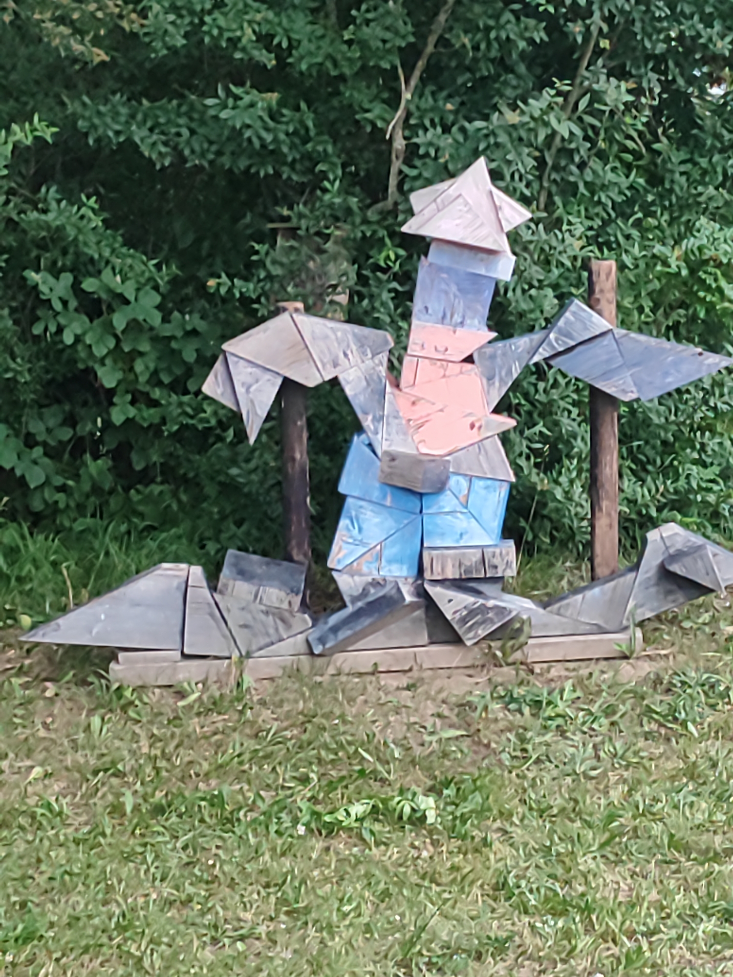 The image shows a unique wooden sculpture in Diedorf, Germany. The sculpture is abstract and appears to be made from various geometric wooden pieces assembled together. It features a central figure with outstretched "wings" and a pointed headpiece, resembling a stylized bird or a whimsical character. The sculpture is painted in muted colors, with shades of grey, blue, and a touch of pink on the central body. It is mounted on a base of wooden planks and is situated outdoors in a grassy area with a backdrop of dense green foliage, likely trees and bushes. The lighting suggests it is daytime, possibly overcast, as there are no strong shadows. The sculpture is the focal point of the image, with the natural environment serving as its setting.