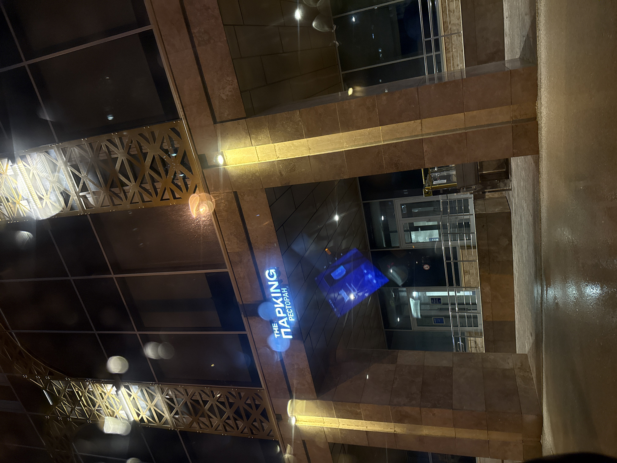 The image displays the exterior of a modern building at night, oriented vertically on its side. The architecture features a facade primarily constructed from light brown and beige-toned stone or tile panels, interspersed with large, dark reflective glass windows.
A prominent illuminated sign is visible on the building, displaying the English text "THE PARKING" and the Cyrillic word "РЕСТОРАН" (RESTORAN), rendered in bright white capital letters, indicating a restaurant. Below this sign, a large, rectangular blue reflection or screen is visible on the glass.
A notable decorative element is a golden-colored metal railing or grille with an intricate, repeating geometric pattern, possibly serving as a balustrade or a decorative architectural accent. Several artificial lights are mounted on the building's facade, casting bright, warm glows and creating lens flares across the dark glass surfaces.
The ground or pavement visible in the foreground and to the right appears wet and highly reflective, suggesting recent rain or damp conditions. The overall ambiance is dark, illuminated solely by artificial lights emanating from the building and street, characteristic of an urban night scene. No people are visible in the frame. The presence of Cyrillic text explicitly points to the location context of Perm, Russia.