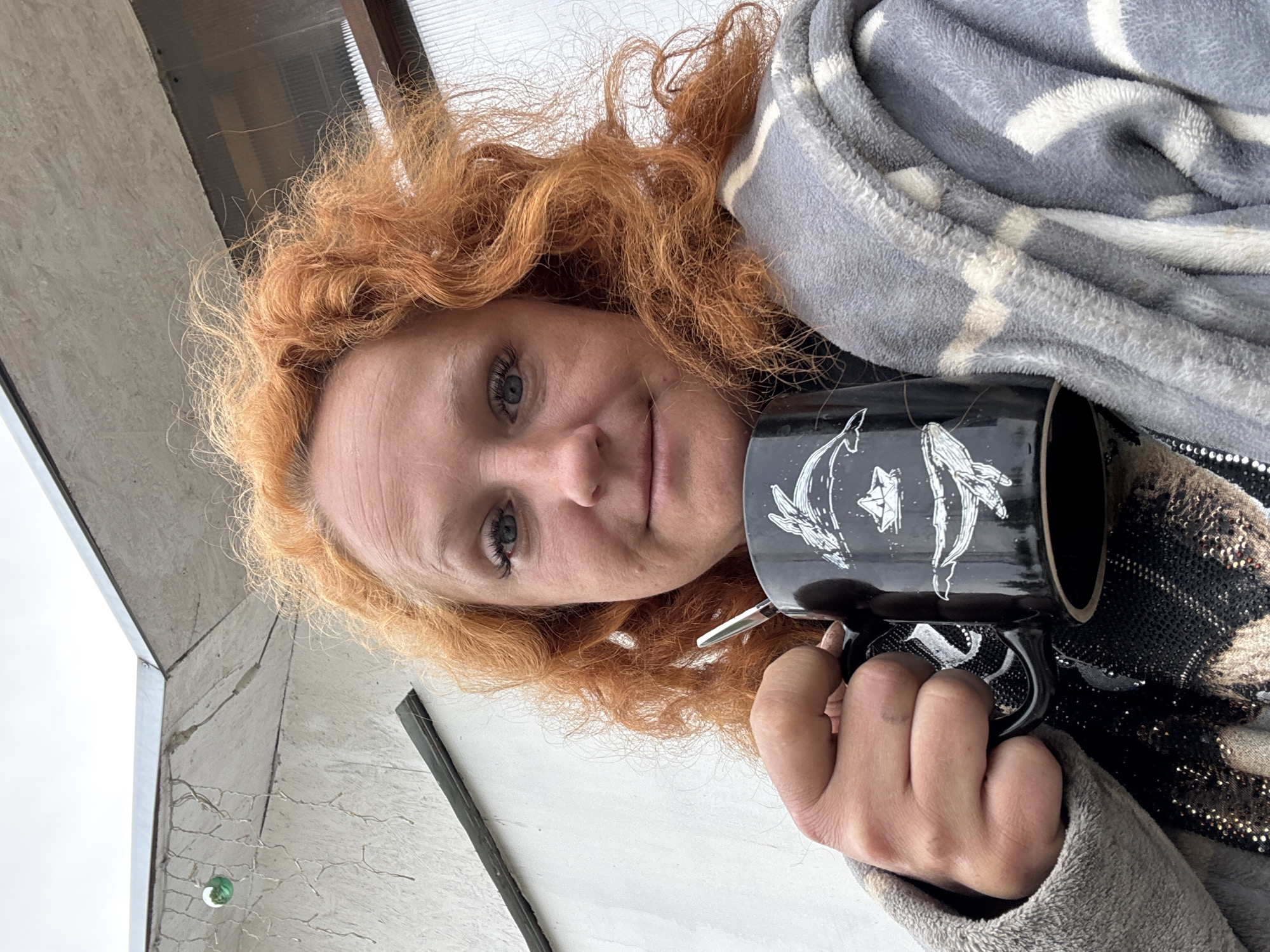 A low-angle shot captures a red-haired woman looking directly at the camera with a pleasant, slightly smiling expression. Her voluminous, curly red hair frames her face. She is holding a black mug adorned with white, stylized illustrations of what appear to be whales or dolphins, possibly intertwined with celestial motifs.
She is draped in a soft, gray blanket with white striped patterns, and a glimpse of a black and gold sequined garment is visible beneath it. The background features a light-colored building structure, possibly concrete or stucco, with a window pane visible in the upper portion. Below this, against the building, there are thin, tangled white wires, resembling fairy lights, with one distinct green spherical ornament. The sky is bright and overcast, indicating daytime under cloudy conditions. The scene suggests a relaxed moment outdoors, possibly on a balcony or near a building in Chaikovsky, Russia. There is no visible text in the image.