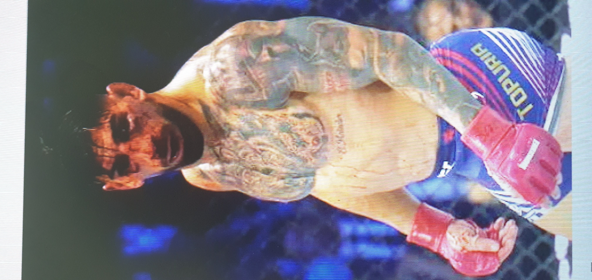 The image depicts a male fighter in a close-up shot, likely during a combat sports event. He is wearing red MMA gloves on both hands. His body is heavily tattooed, with intricate designs visible on his torso and arms. The fighter is looking towards the left of the frame, with his mouth slightly open, potentially mid-action or during a break. His expression is intense, suggesting focus or exertion.
He is wearing blue shorts with red and white stripes, and the word "TOPURIA" is prominently displayed in white lettering on the shorts, likely indicating his name or a sponsorship. The background is dark and blurred, with hints of a blue, grid-like structure that could be a cage or netting, typical of an MMA arena. The lighting is dramatic, highlighting the fighter and his tattoos. The image appears to be a still from a video recording, as evidenced by the horizontal scan lines present throughout the frame, suggesting it was captured on a screen. There is no indication of the time of day or weather conditions, but the setting is clearly an indoor sporting venue.