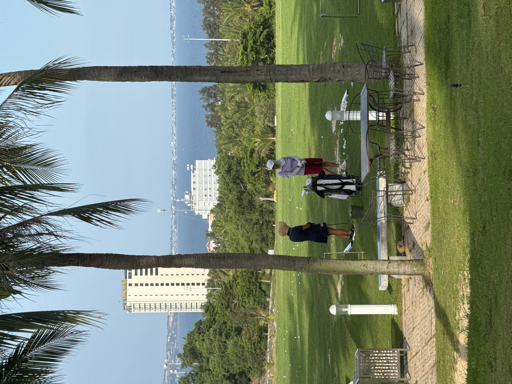 The image depicts a bright, sunny day at a golf driving range in Phu Thuy, Vietnam, with the ocean and a high-rise building visible in the background. Two individuals, presumably golfers, are present on the course. One, wearing a straw hat and a dark blue dress, stands to the left, holding a golf club. The other, dressed in a light purple shirt and red shorts, stands to the right, also near a golf bag. Numerous white golf balls are scattered across the well-maintained green grass. In the foreground, a paved area features a metal table and chairs, as well as a stone bench and a trash receptacle. Palm trees frame the scene, casting shadows on the ground. The overall atmosphere suggests a recreational outing in a coastal environment.
