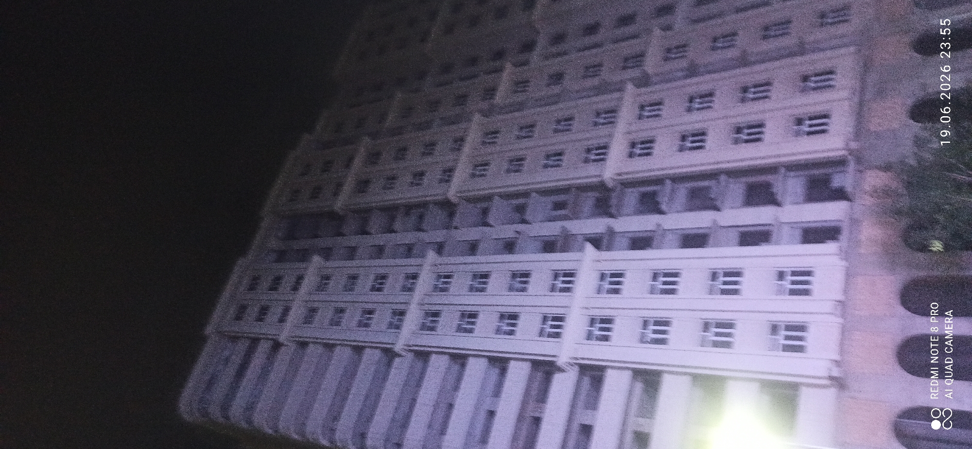 An outdoor, low-angle, night-time photograph captures a tall, light-colored apartment building. The building features a repeating pattern of windows and balconies, with vertical decorative elements dividing sections of the facade. The sky above is dark and featureless. The bottom left corner of the image contains a watermark that reads "REDMI NOTE 8 PRO AI QUAD CAMERA," and the bottom right corner displays a timestamp "19.06.2026 23:55." A faint light source is visible on the left side of the building. The base of the building appears to have arched openings.