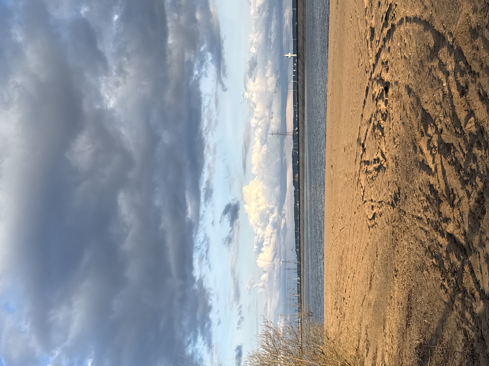 The image displays a vast landscape scene, primarily featuring a dynamic sky, a body of water, and a sandy shoreline, captured during what appears to be late afternoon or early evening.
The upper half of the frame is dominated by a dramatic sky, characterized by a mix of heavy, dark gray clouds on the left and brightly illuminated, white cumulus clouds on the right. Patches of pale blue sky are visible in between, especially towards the horizon.
A wide, calm body of water spans the mid-ground, acting as a clear division between the sky and the foreground. The water reflects the subdued light from the sky, appearing as a smooth, grey-blue expanse. Along the distant horizon line, a series of human-made structures are visible, including several tall transmission towers or power lines. A distinct, white, beacon-like structure stands out against the brighter clouds, possibly a communication tower or a navigational aid.
The lower half of the image features a sandy or dirt bank, rich with texture. It shows numerous imprints, likely a combination of footprints and tire tracks, indicating recent activity. The warm, low-angle sunlight casts gentle shadows and highlights, emphasizing the contours and irregularities of the sand. In the very bottom right corner, sparse, dry vegetation, resembling light brown brush or reeds, is visible.
There are no people or visible text in the scene. The overall impression is one of expansive, natural beauty with a touch of distant human presence. The lighting suggests a tranquil, contemplative atmosphere. Given the context, this scene is likely a shore of a large lake or reservoir in the Noviy, Russia area.