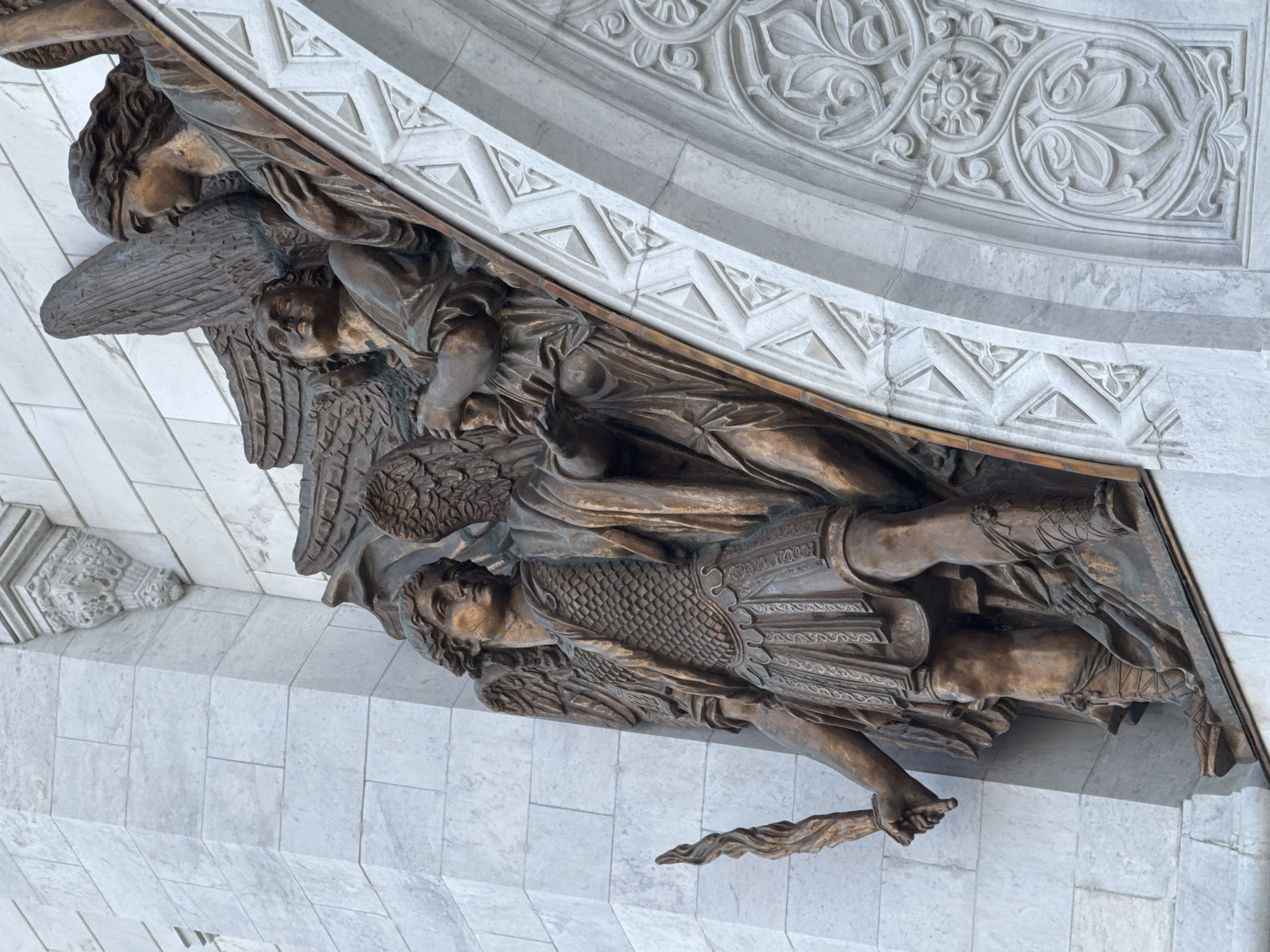 The image depicts a bronze sculpture of the Archangel Michael with other angels, situated on the facade of the Cathedral of Christ the Saviour in Moscow, Russia. Michael is in armor with a sword and wings. The sculpture is mounted on a stone structure adorned with geometric and floral designs.
