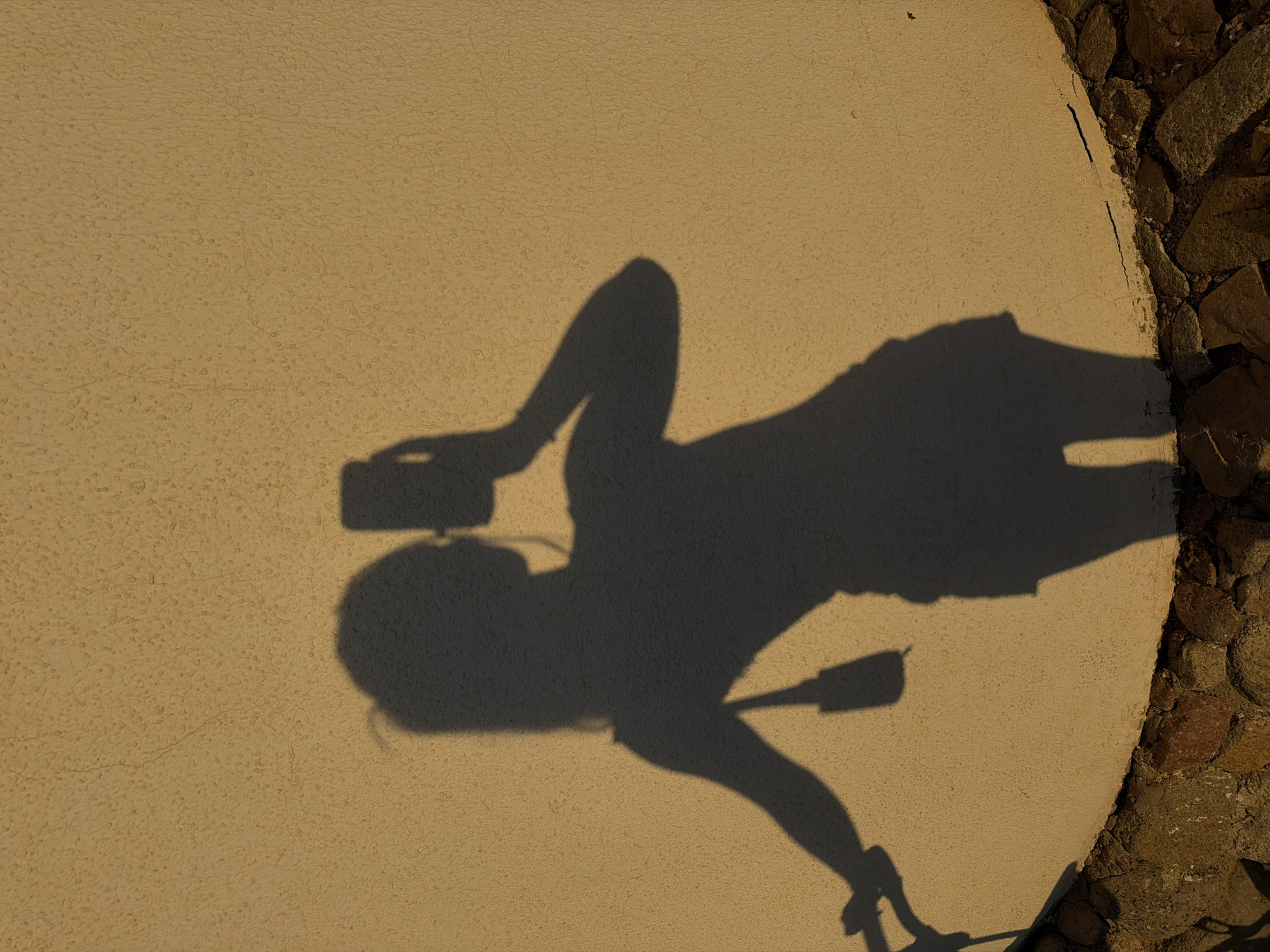 This image shows the shadow of a person taking a selfie with a smartphone. The person appears to be standing, with one arm raised to hold the phone, and the other arm bent at the elbow. The shadow reveals the person is wearing shorts and possibly a t-shirt. The background is a textured, light-colored wall, with a circular section of rocks on the right side. The strong shadow indicates a bright, sunny day, likely in the late afternoon or early morning given the angle of the light. The setting appears to be outdoors, possibly a patio or courtyard, with the rough texture of the wall and the natural stones suggesting a warm, possibly arid climate like Sharm El Sheikh, Egypt. There is no visible text in the image. The overall mood is casual and summery.