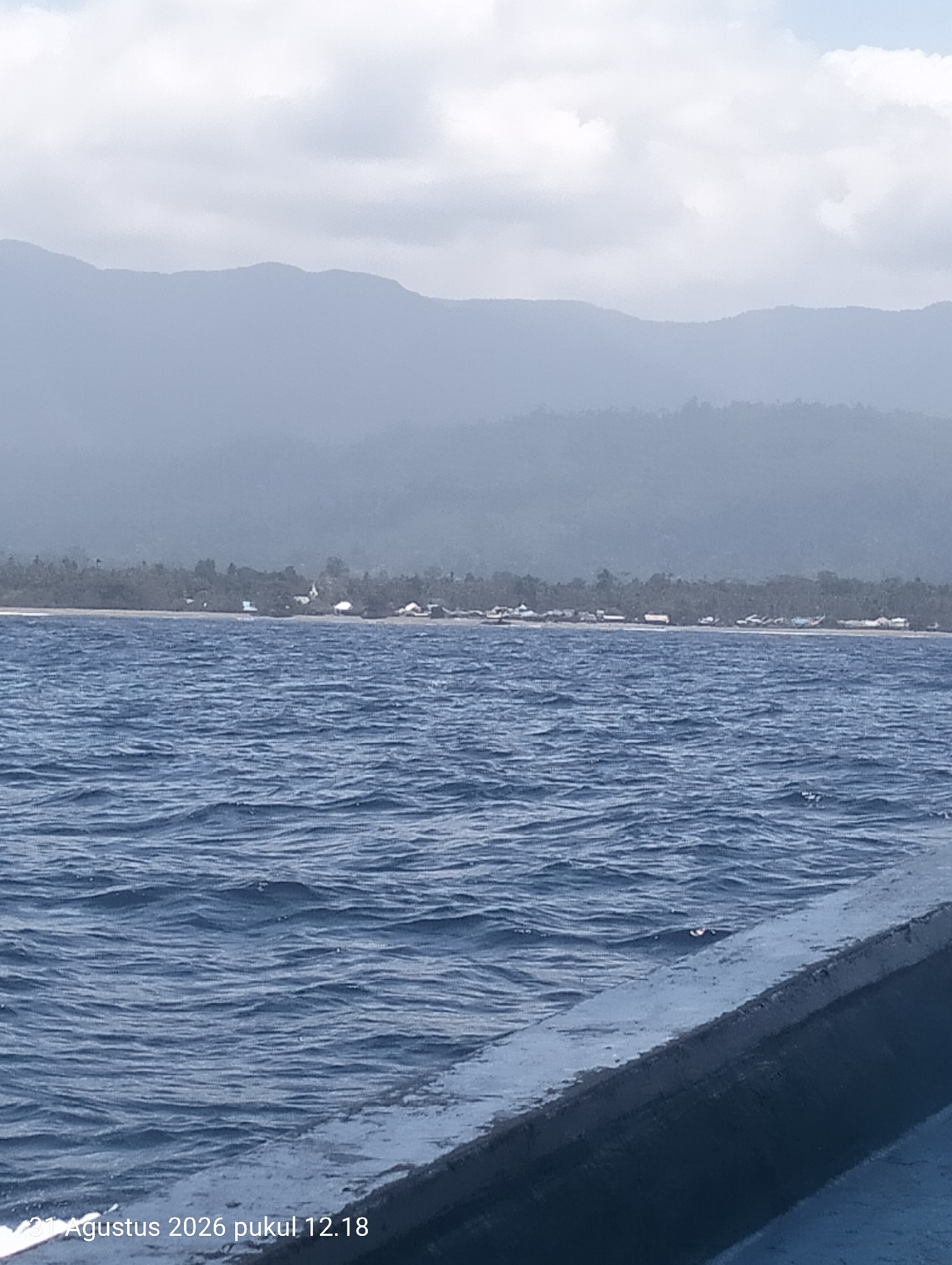 The image depicts a body of water with gentle waves, likely the sea, in the foreground. The water is a deep blue color. A dark, curved object, possibly the edge of a boat or vessel, occupies the bottom right portion of the frame. In the middle ground, a sandy beach is visible, lined with a dense growth of trees and scattered small buildings. This settlement appears to be located on the coast. In the background, a range of misty, blue mountains rises, their peaks softened by atmospheric haze. The sky above is a pale blue with scattered white clouds. A timestamp in the bottom left corner reads "31 Agustus 2026 pukul 12.18", indicating the date and time the image was captured. While the location is stated to be Pihingtado, Indonesia, this information cannot be independently verified from the visual content of the image itself. There are no people or discernible actions depicted in the scene.