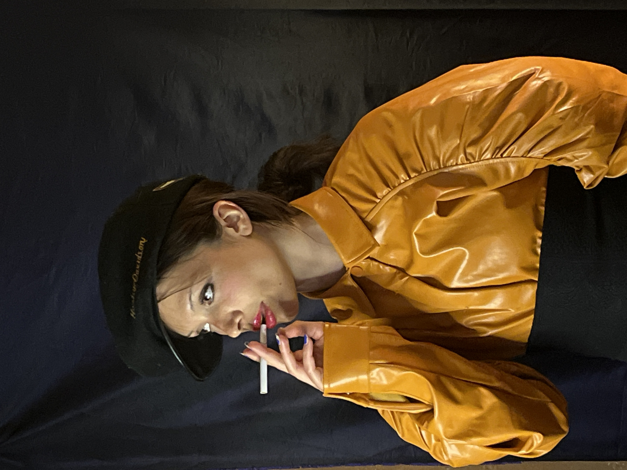 A woman with fair skin and dark hair, wearing a black cap with gold lettering, a bright yellow leather jacket, and red lipstick, is holding a cigarette in her right hand. She is positioned against a dark blue or black background. The location cannot be determined from the image.