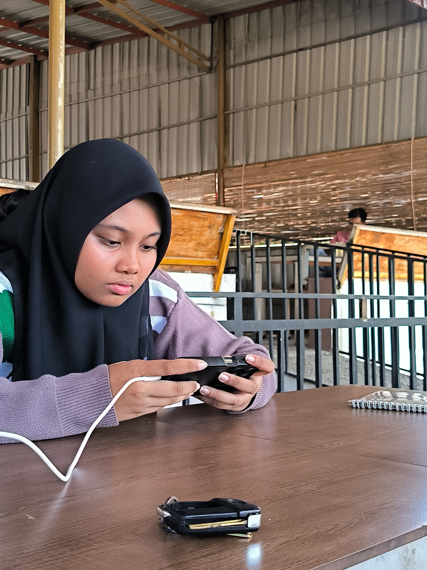 A woman wearing a black hijab and a lavender sweater is sitting at a wooden table, looking at a black smartphone that is connected to a white charging cable. On the table is a black car key fob and a spiral-bound notebook. The woman is in a covered area with a corrugated metal roof supported by metal beams. In the background, a metal railing is visible, and a person is partially visible standing behind the railing on a higher level. The setting appears to be a public or semi-public space, possibly in Matang, Indonesia.