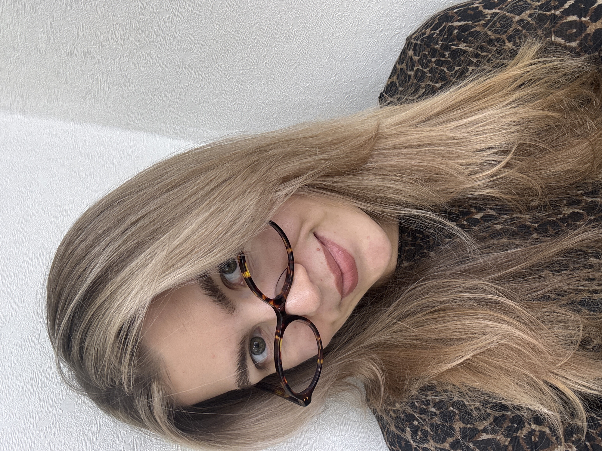 The original image displays a close-up portrait of a person, likely a woman, with their head tilted significantly to the left (or the image rotated 90 degrees clockwise). The subject is looking upwards directly at the viewer with a subtle, friendly smile. They have long, flowing light blonde hair with varying shades, which frames their face and extends across the frame. Large, round glasses with a prominent tortoiseshell pattern are perched on their nose, framing their greenish-grey eyes. The person has full eyebrows and their lips are slightly parted, revealing a hint of tongue and a natural lip color with a soft sheen. They are wearing a black and brown leopard print garment, which is visible on the right side and bottom of the frame. The background consists of a plain white wall with a textured surface, suggesting an indoor setting. The lighting is soft and even, illuminating the subject's face without harsh shadows. No text is visible in the image.
