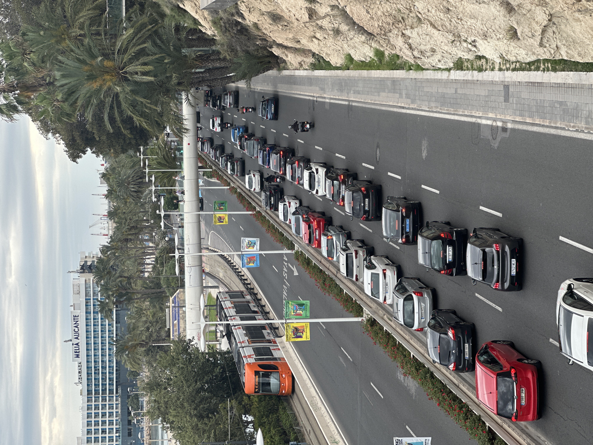 The image captures an aerial view of a road in Unknown, Unknown. The road features a line of vehicles, including cars and a motorcycle, traveling in the same direction. Alongside the road, there is a tram. The Melia Alicante building can be seen in the distance. The environment includes trees, a cliff face, and a paved area.