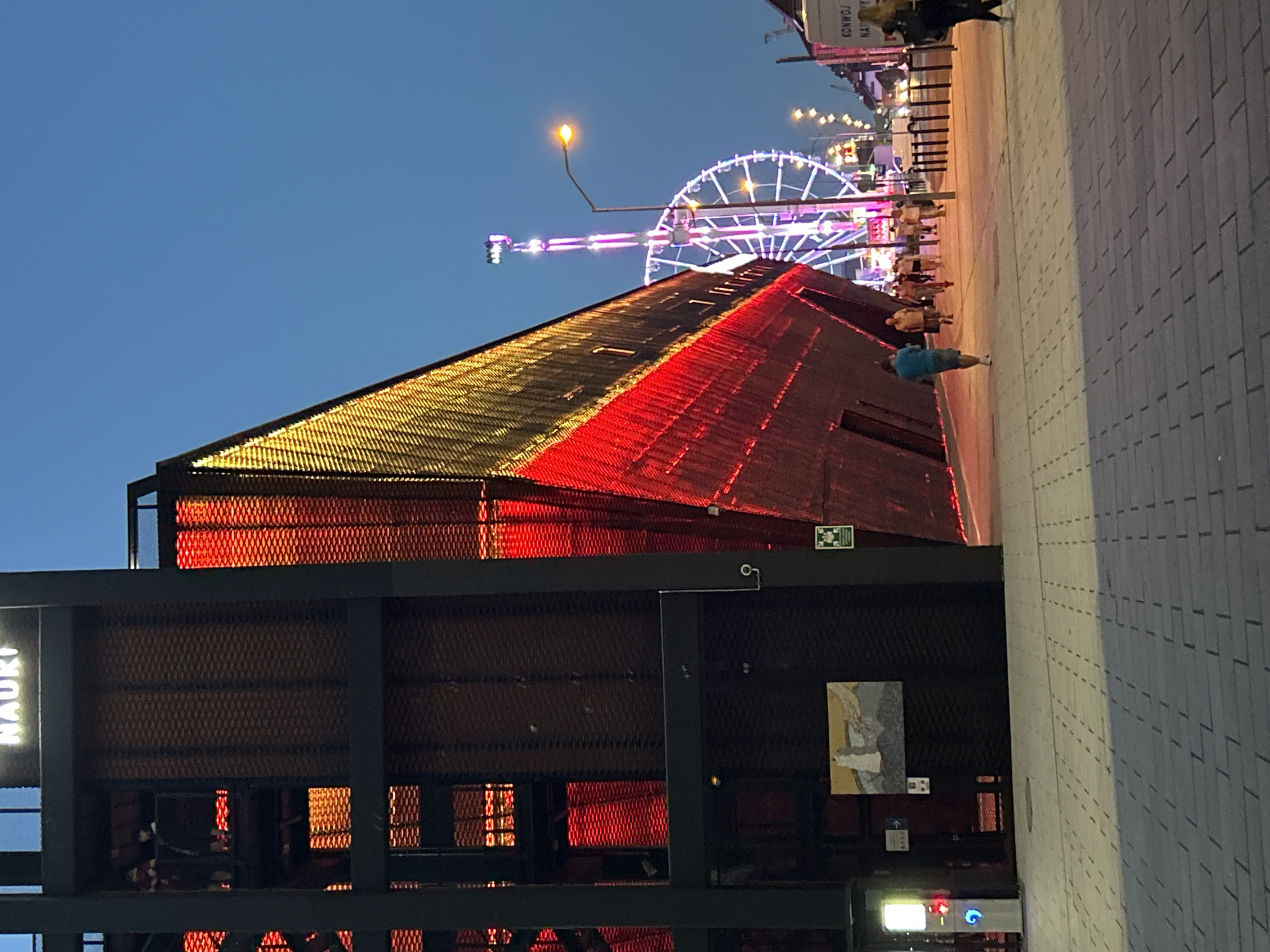 The image captures a scene at dusk in Szczecin, Poland, specifically at the "Centrum Nauki" (Science Center). The prominent structure on the left is the modern building of the Science Center, characterized by its black metallic framework and a striking facade illuminated with red and yellow lights, designed to resemble a ship. Several people are visible on a walkway in front of the building. In the background, a brightly lit Ferris wheel and a tall amusement ride illuminate the twilight sky. The ground is paved with brick-like tiles. The overall atmosphere suggests a lively evening in an urban waterfront area.