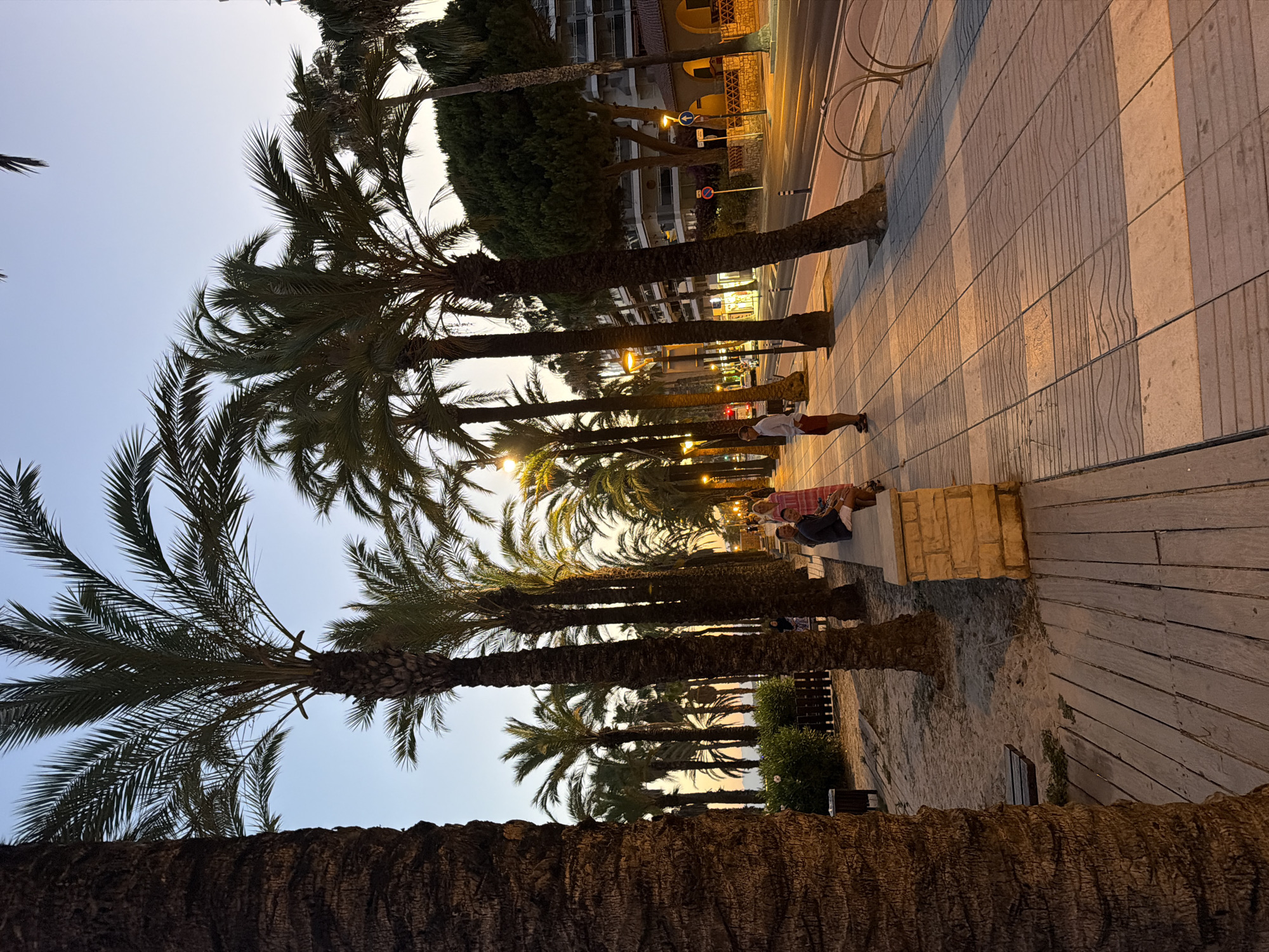 In Salou, Spain, on a waterfront promenade lined with palm trees, the late afternoon sun casts long shadows across the tiled walkway. A group of people are visible: a couple in the foreground, with a woman in a patterned dress and a man in shorts and a t-shirt, are seated on a low stone wall. Further down the promenade, a man in a white shirt and red shorts stands looking towards the street. Across the street, buildings with balconies are lit up, and streetlights are beginning to illuminate the scene. The sandy area beside the promenade has wooden planks and benches, and trash bins are present. The sky is a gradient of pale blue and orange, indicating sunset.