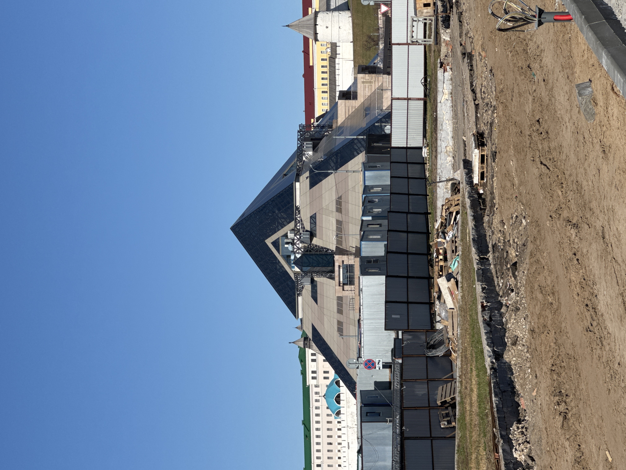 The image depicts a construction site in Kazan, Russia, dominated by the pyramid-shaped Kazan National Cultural Center. The building features a dark glass pyramid structure atop a lower, wider base constructed with lighter-colored materials. Scaffolding and other construction elements are visible on the building.
The ground is uneven and composed of dirt and gravel. A series of dark, rectangular structures resembling storage containers or temporary offices run along the perimeter of the construction site. Behind these, a variety of buildings, including older structures with architectural details such as turrets and a tower with a spire, are visible. The sky is clear and blue, indicating a bright day.