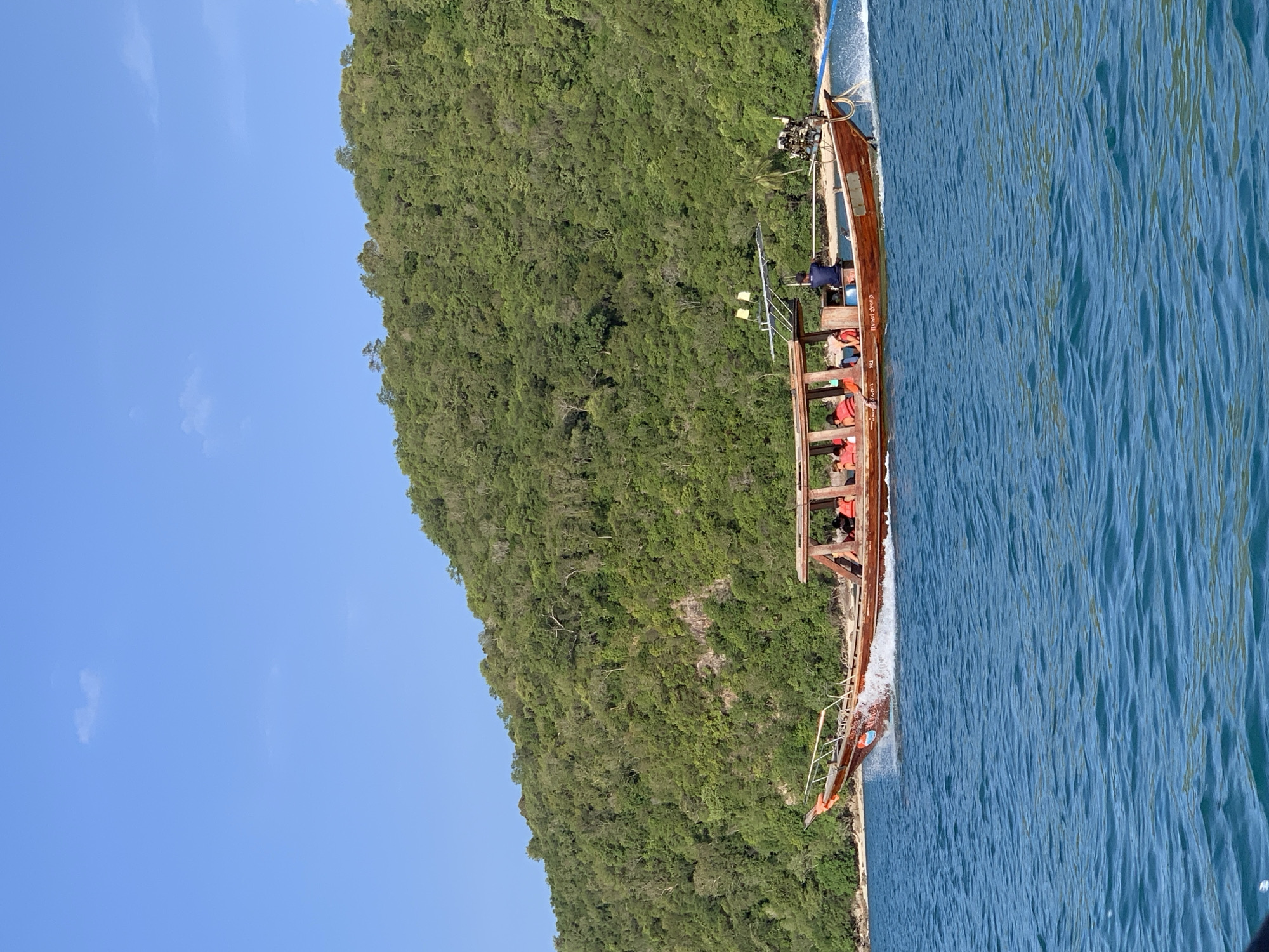 In this medium shot, a wooden boat travels across a bright blue body of water. The boat is long and narrow with an open cabin area covered by a canopy. Several passengers, wearing life vests, sit inside the cabin. A man stands at the stern of the boat, steering. The boat creates a white wake behind it. In the background, a lush green, tree-covered island slopes down to the water. The sky above is clear blue with a few small, wispy clouds. The scene appears to be a tourist location in Thailand.