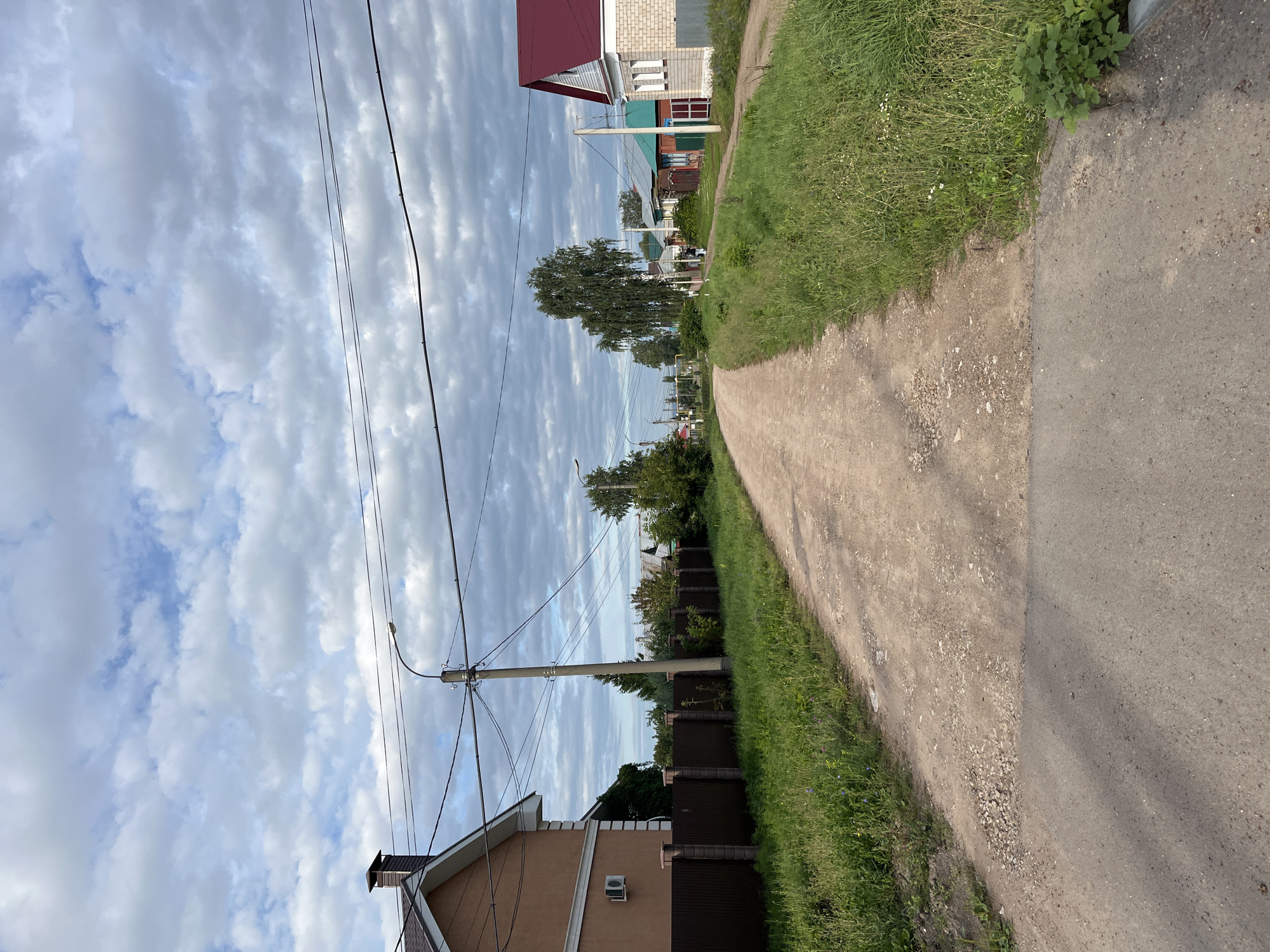The image depicts a view down a dirt road in a rural or suburban area, likely a village or dacha settlement, with houses lining both sides of the road. The sky is partially cloudy, with a mix of blue sky and white, fluffy clouds. The road itself is unpaved and appears to be in fair condition, with some patches of loose gravel and small potholes. On the left side of the road, there is a brown fence and the side of a house with an air conditioning unit attached. On the right side, there are several houses with various colored roofs and brick and painted facades. Green grass and trees are abundant on both sides of the road, indicating a natural, undeveloped environment. Power lines are visible crisscrossing the sky, supported by utility poles. In the distance, the road curves to the left, and there are more houses and trees visible. A single person is walking away from the camera down the road. The overall impression is of a quiet, residential area in a natural setting. It is not possible to definitively confirm the location as Ufa, Russia, without further information.