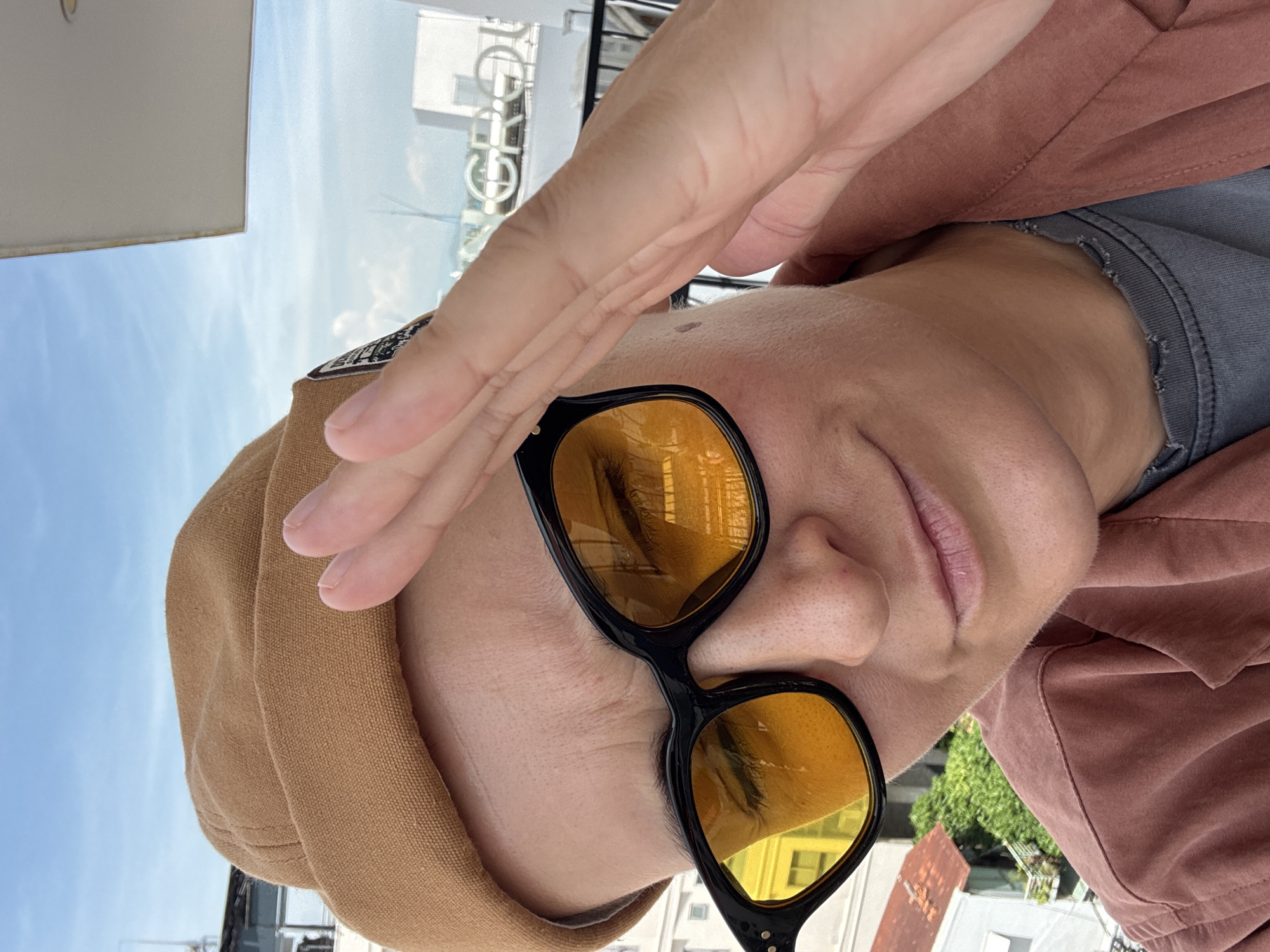 A person, wearing a tan beanie, black-rimmed sunglasses with yellow lenses, and a rust-colored jacket over a gray t-shirt, poses for a selfie on a balcony overlooking a cityscape. The person's right hand is raised, shielding their face from the sun. In the background, buildings and lush greenery are visible, with a glimpse of the sky above. The scene appears to be in Nha Trang Ward, Vietnam.