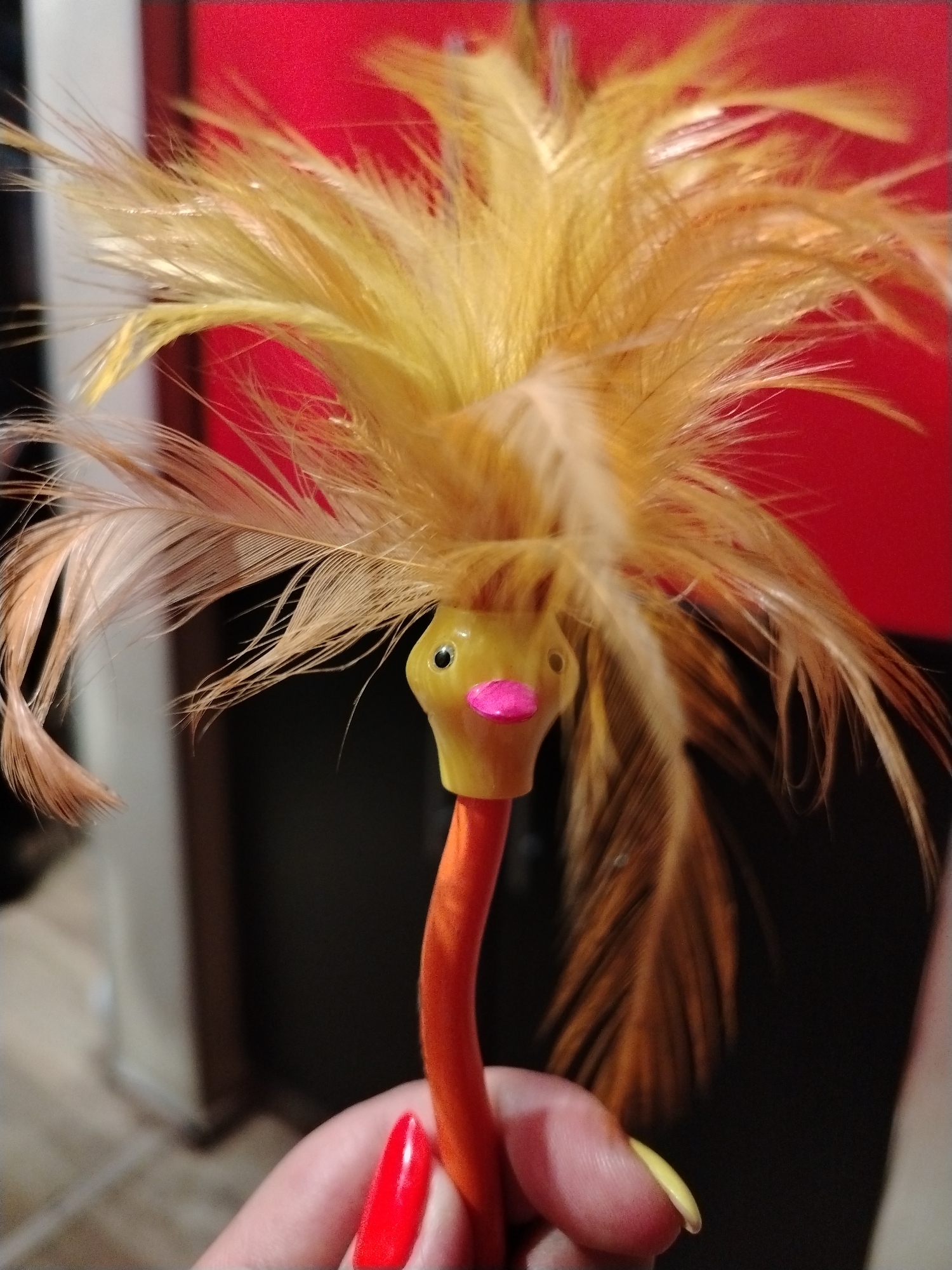 A hand holds a pen in the shape of a yellow bird with feathers on its head. The bird has a pink beak and black eyes. The hand has red nail polish on one finger and yellow nail polish on another. A red wall can be seen in the background. The location is presumed to be Chaikovsky, Russia, based on the prompt.