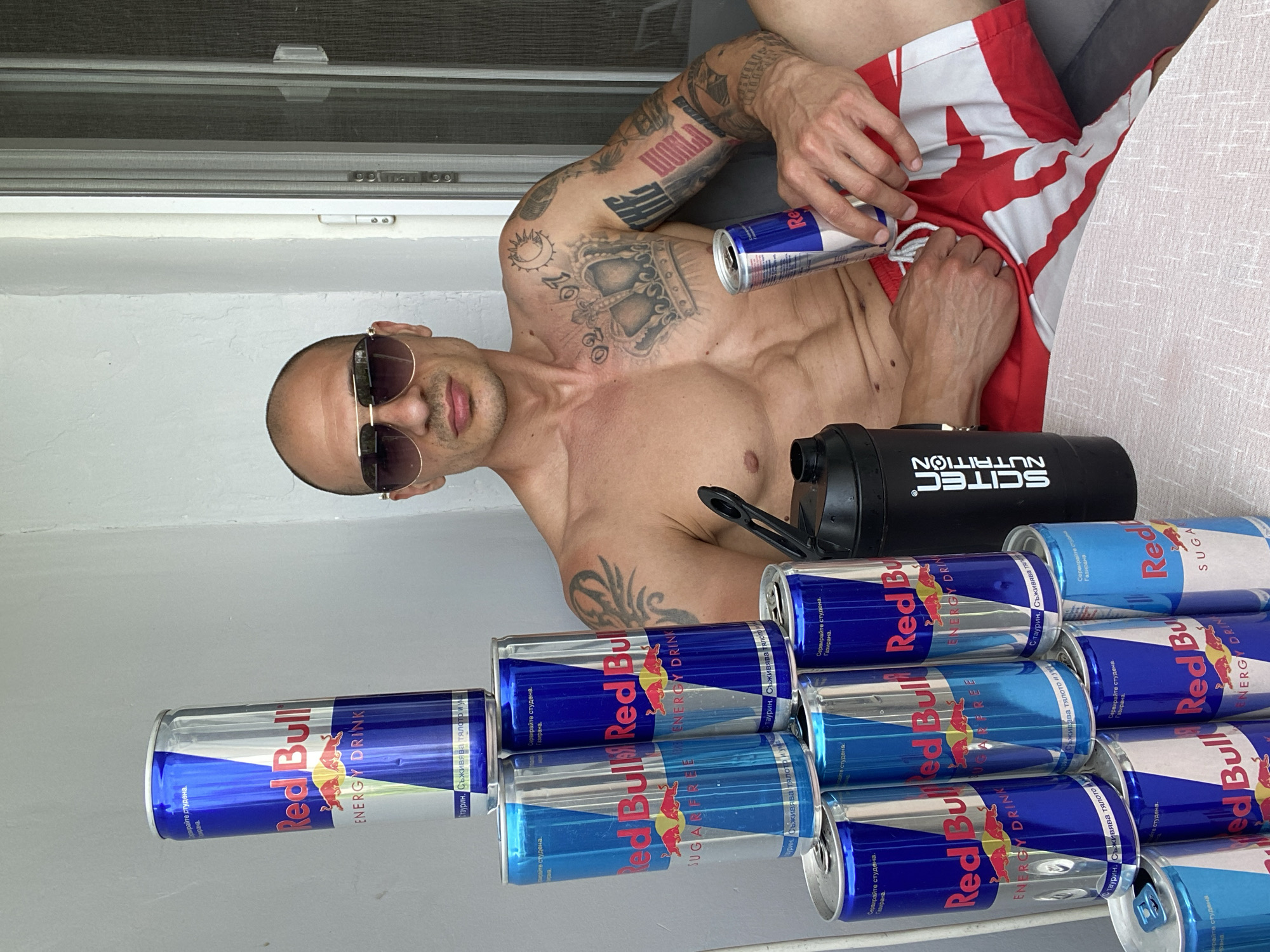 A shirtless, bald man with sunglasses and numerous tattoos sits on a couch next to a tower of Red Bull cans and a black shaker bottle. He is holding a Red Bull can in his right hand. The background is a white wall and a sliding door with a screen. The image appears to have been taken in an outdoor setting, possibly a balcony or patio, in Pleven, Bulgaria.